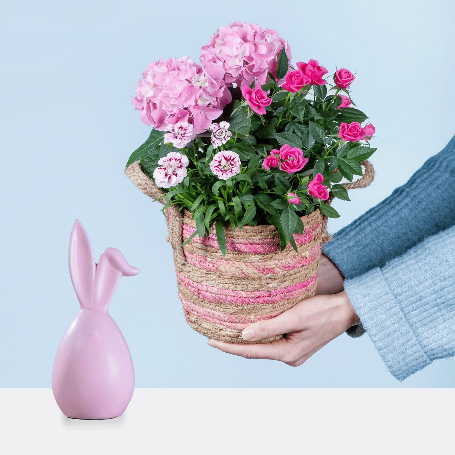 Ein Strauß leuchtend rosa Hortensien und Rosen wird in einem geflochtenen Korb vor einem hellblauen Hintergrund gehalten, im Vordergrund steht eine kleine rosa Keramikvase.