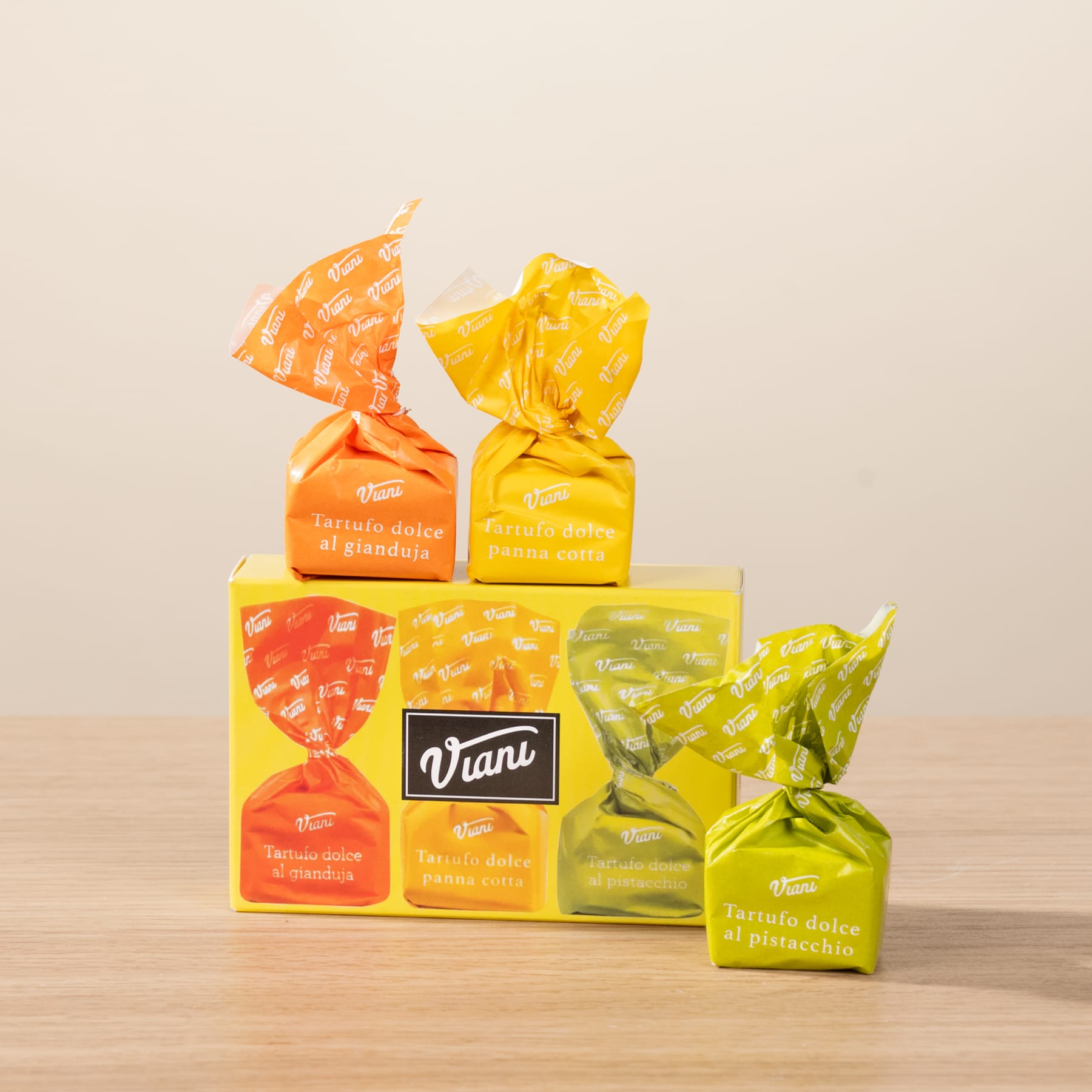 Bunte Packungen mit Snacks der Marke Viano, darunter orange und gelbe Cracker in Dreiecksform, sind auf einer Holzoberfläche vor einem hellen Hintergrund gestapelt.