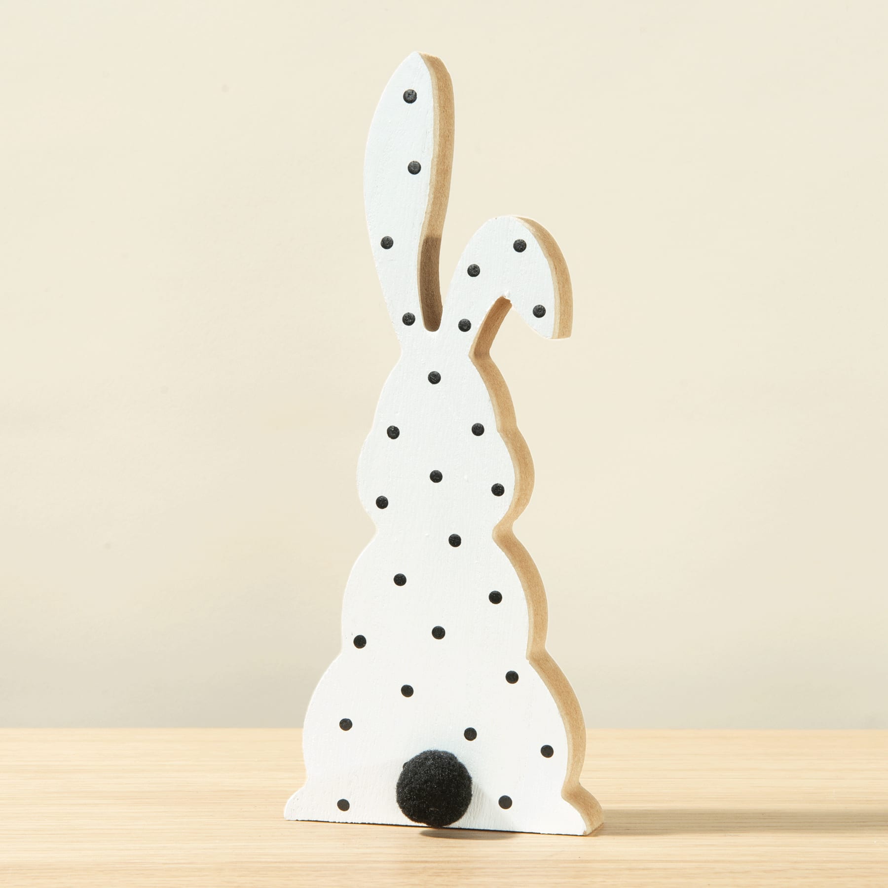 Eine weiße Hasenfigur aus Holz mit schwarzen Punkten steht auf einer Holzfläche vor einem hellen Hintergrund.
