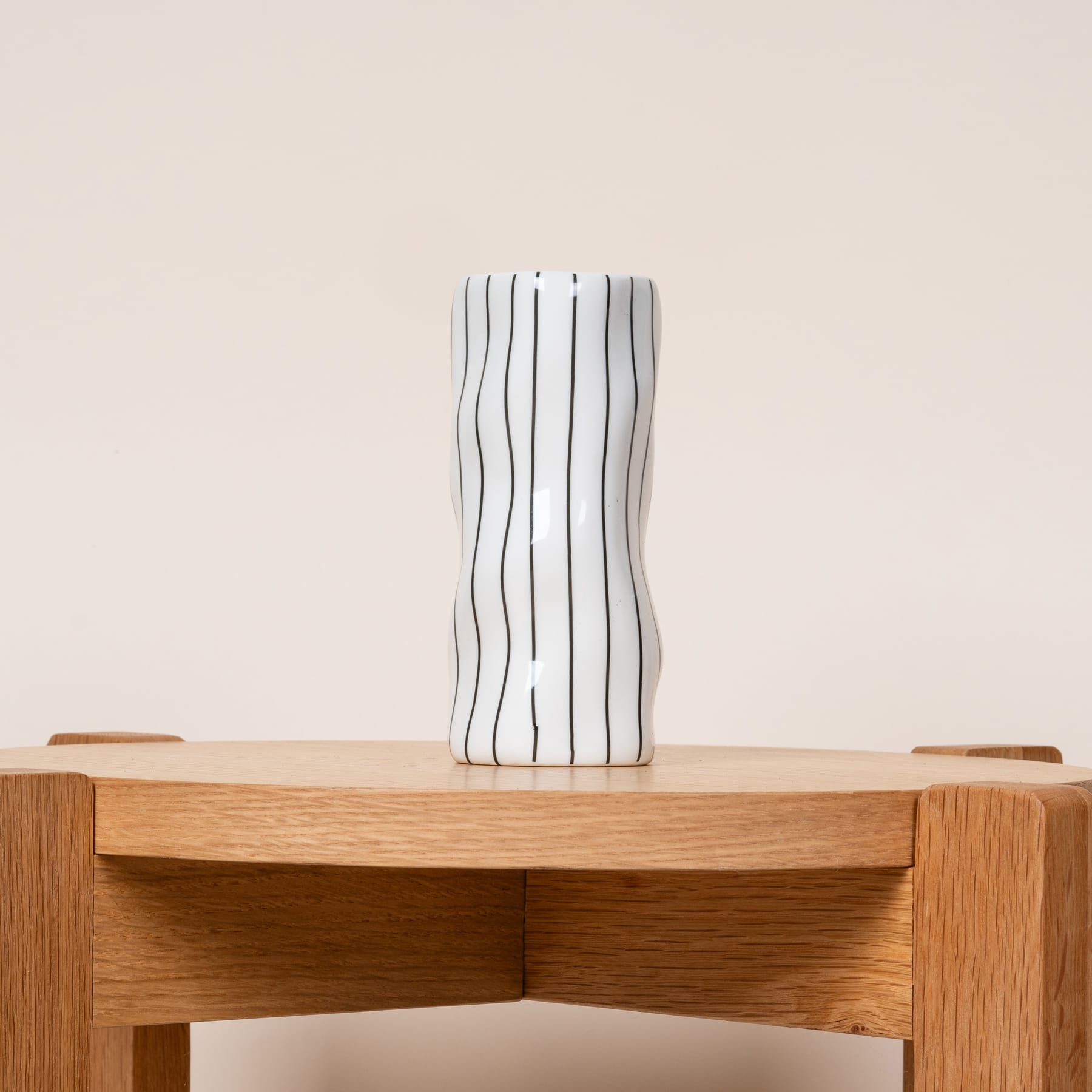 Eine gestreifte Keramikvase steht auf einem Holzregal vor einem schlichten beigen Hintergrund.