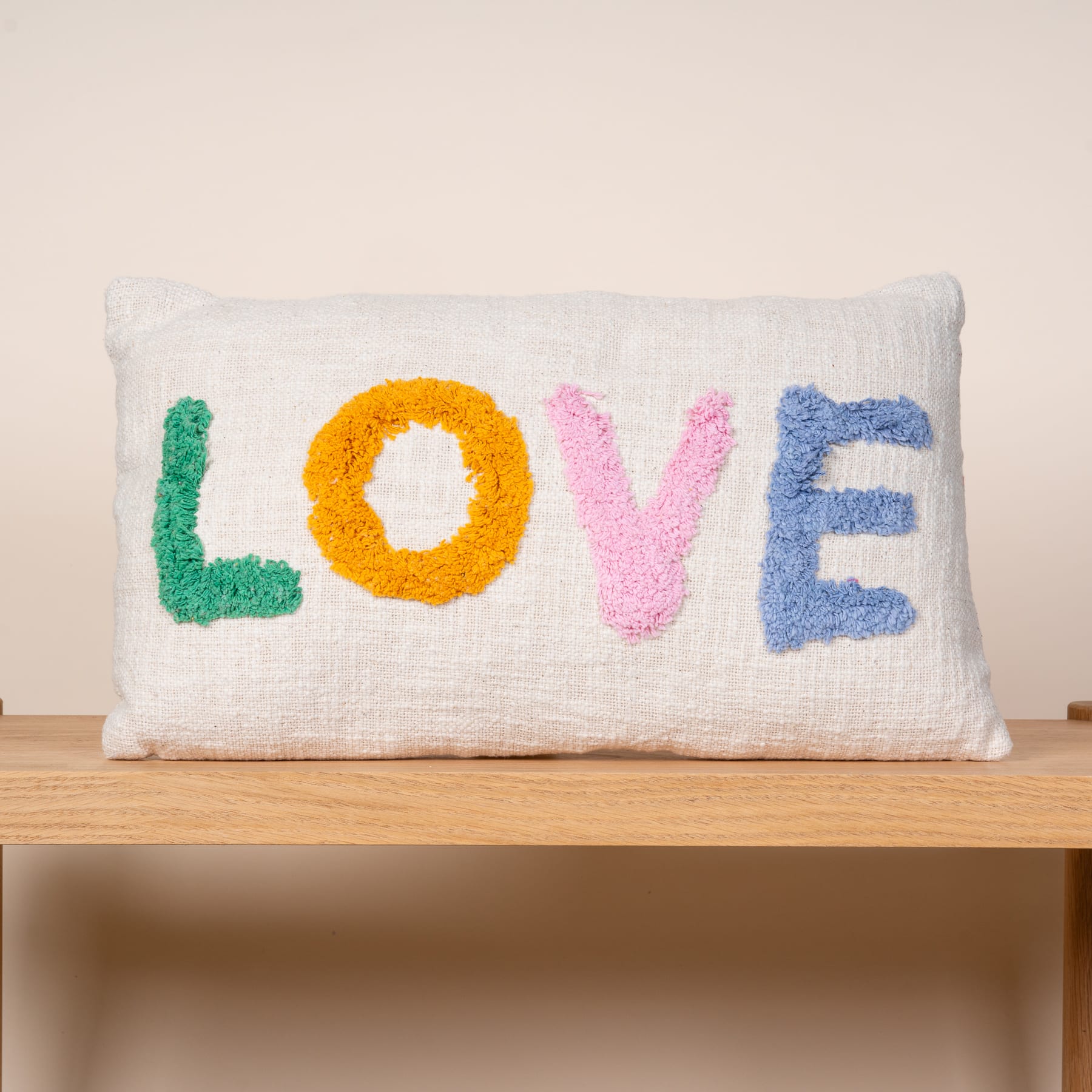 Auf einer Holzoberfläche liegt ein weißes, flauschiges Kissen mit dem Wort „LOVE“ in bunten, strukturierten Buchstaben.