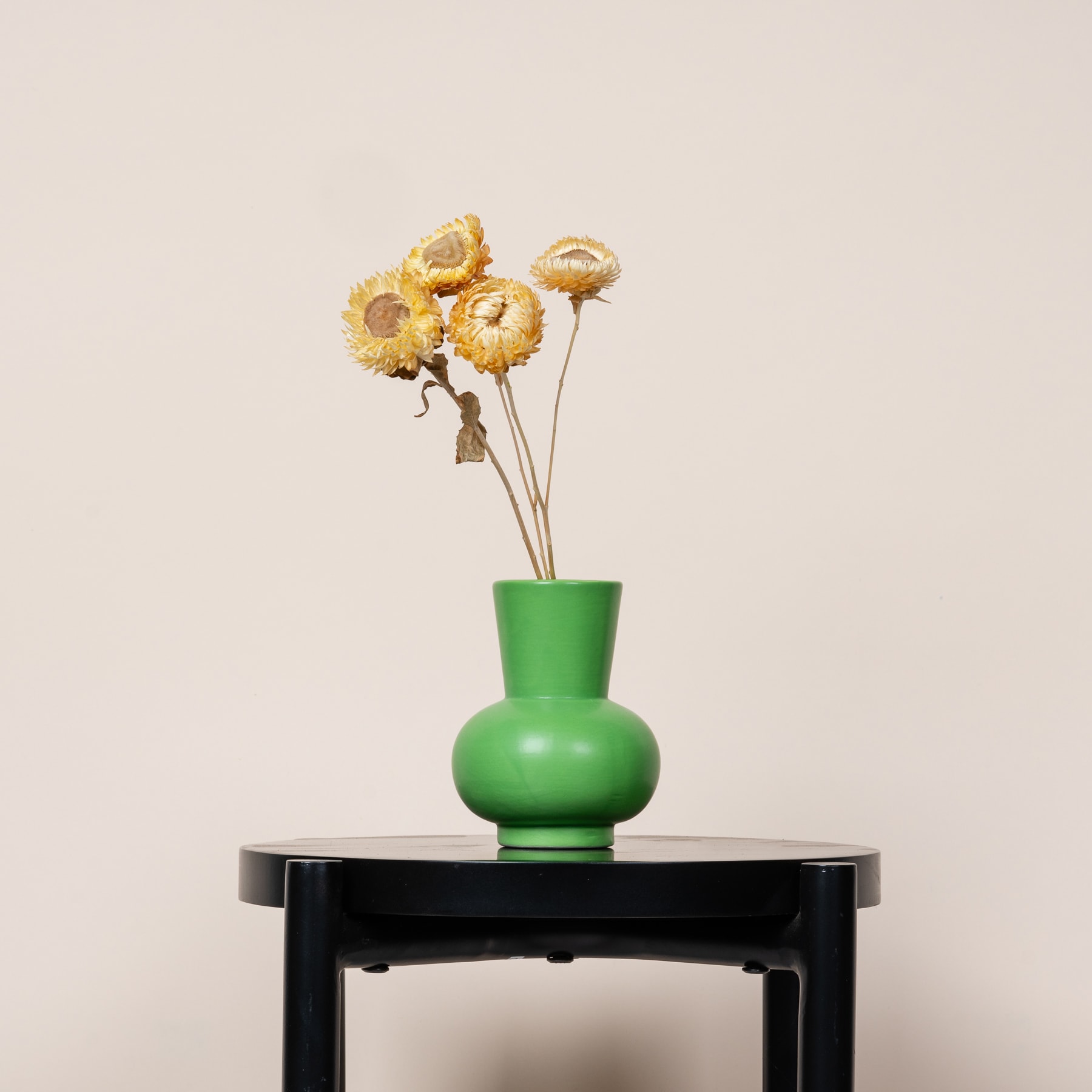 Eine grüne Keramikvase mit einem Strauß getrockneter gelber Blumen steht auf einem schwarzen Holztisch vor einem schlichten beigen Hintergrund.