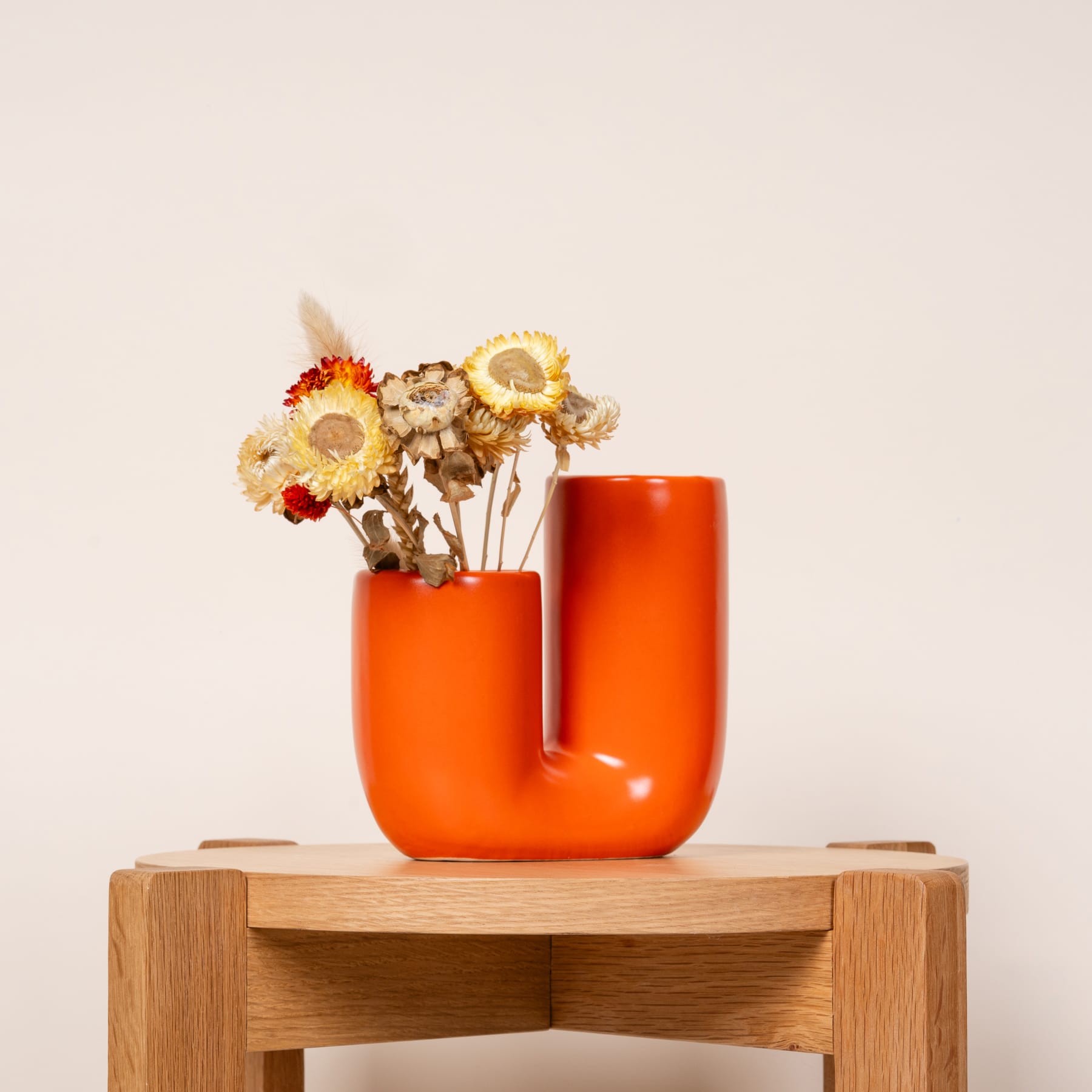 Eine orangefarbene Keramikvase mit einem Strauß getrockneter Blumen steht auf einer Holzplattform vor einem schlichten, hellen Hintergrund.