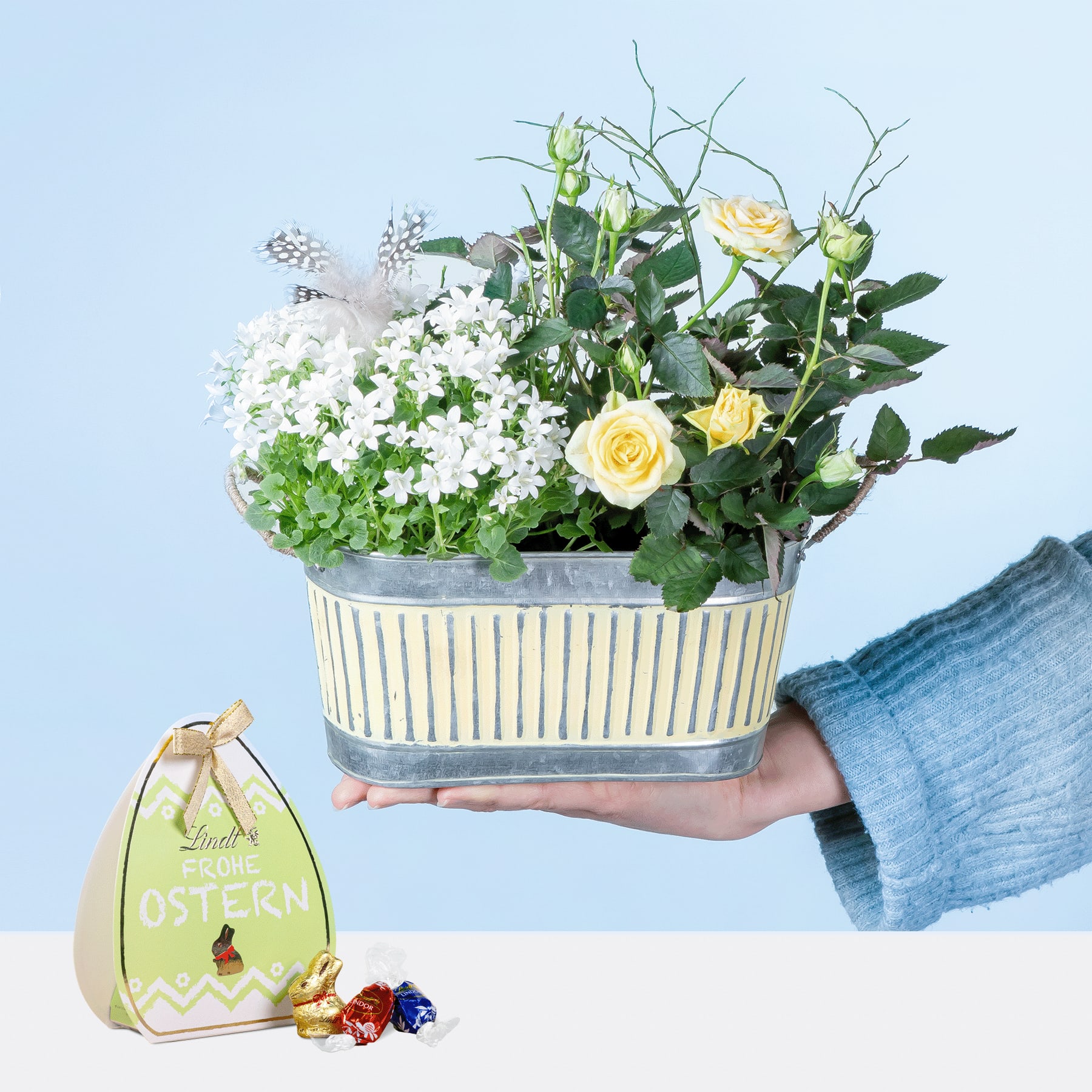 Die Hände einer Person halten einen dekorativen Blumentopf, der mit verschiedenen weißen und gelben Blumen gefüllt ist, mit einer kleinen Dekoration zum Thema Ostern im Vordergrund.