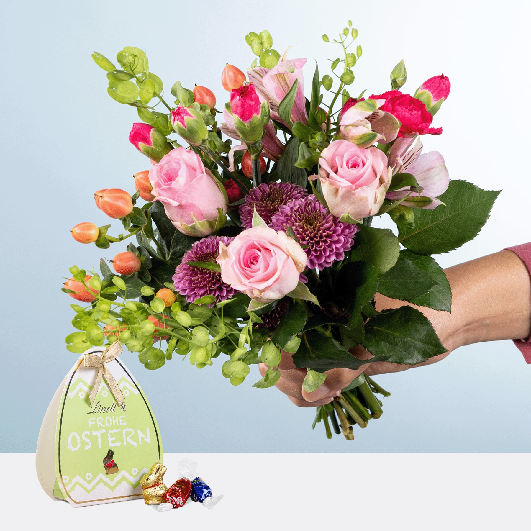 Eine Person hält einen leuchtenden Strauß rosa Rosen, grüne Hortensien und andere bunte Blumen in den Händen, im Vordergrund ist ein kleines Schokoladenei mit Ostermotiv zu sehen.