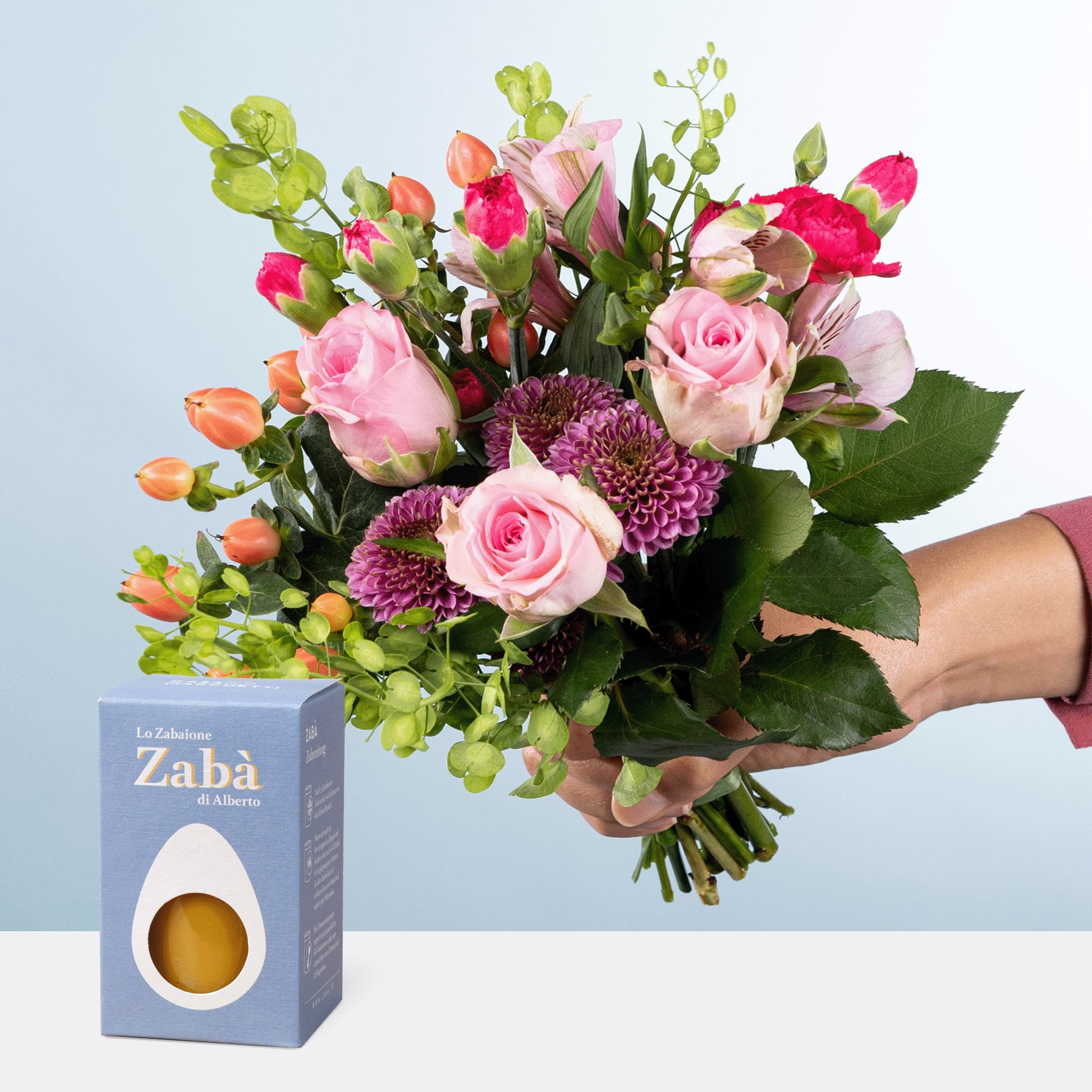 Eine Person hält einen farbenfrohen Strauß rosa Rosen, lila Chrysanthemen und anderen bunten Blumen in den Händen, begleitet von einer kleinen Geschenkbox mit der Aufschrift „Zaba“ im Vordergrund vor einem schlichten blauen Hintergrund.