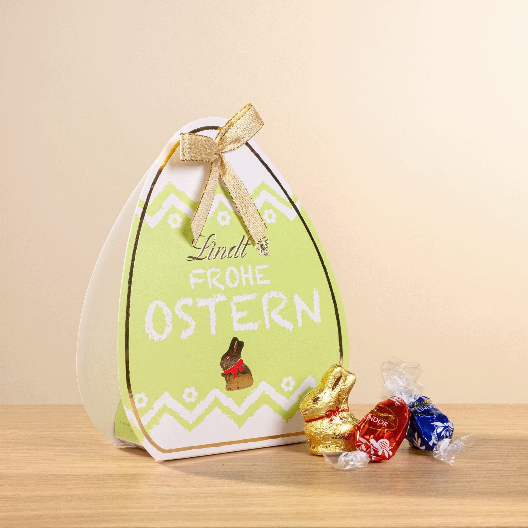 Eine grüne Geschenkbox mit Ostermotiv und einer Schleife darauf steht auf einer Holzoberfläche, umgeben von kleinen Schokoladen-Ostereiern.