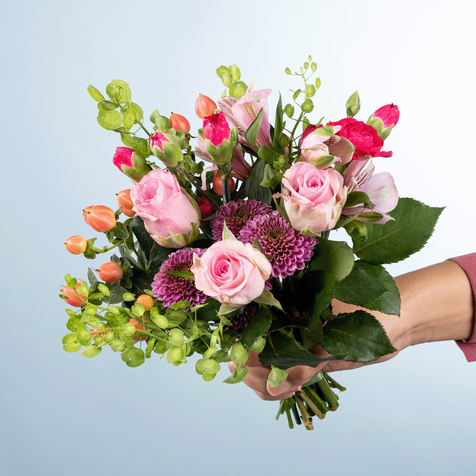 Ein leuchtender Strauß rosa und roter Rosen, Chrysanthemen und anderer Grünpflanzen, den eine Person vor einem schlichten Hintergrund in den Händen hält.