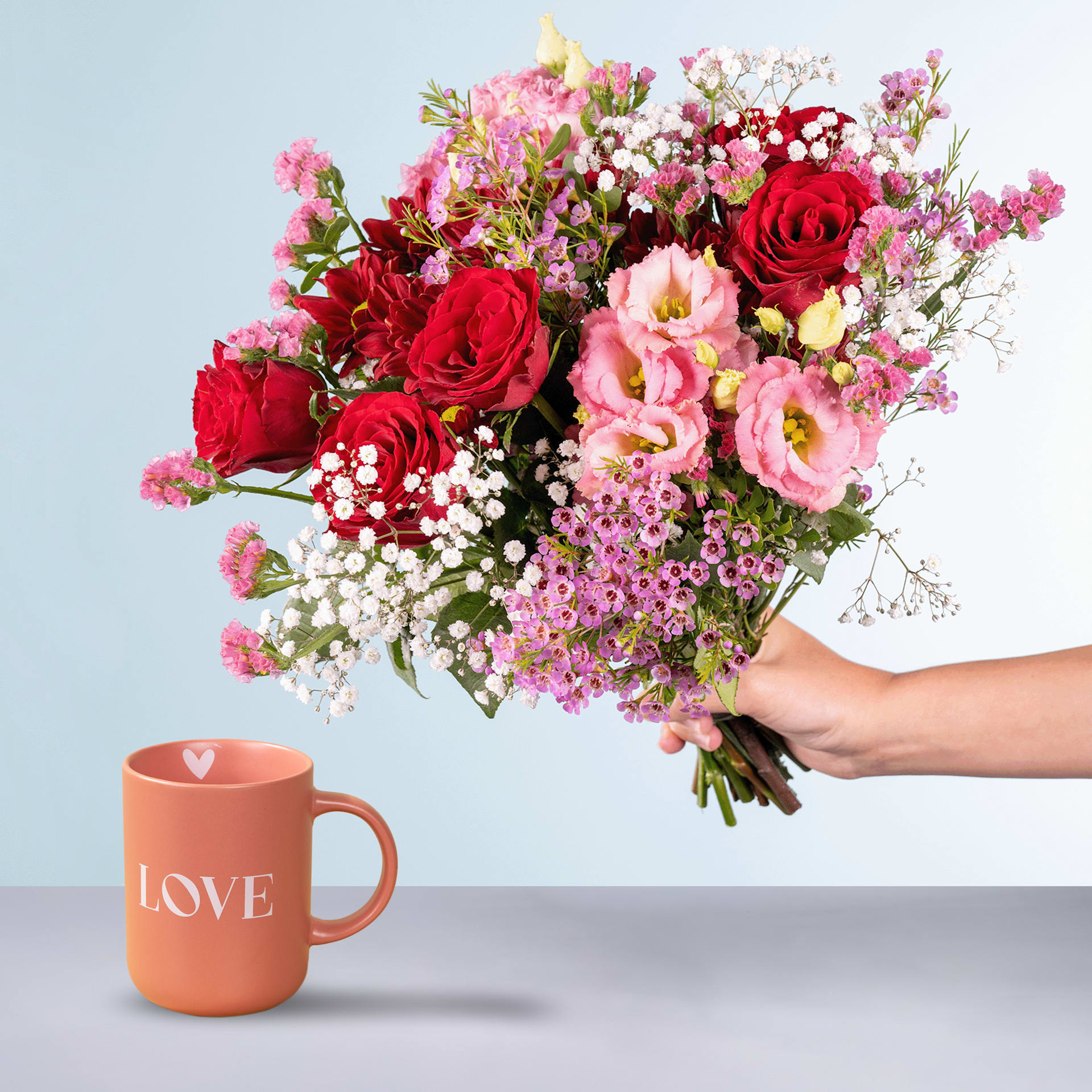 Eine Person hält einen farbenfrohen Strauß roter Rosen, rosa Nelken und zarter Schleierkrautblüten in den Händen. Vor einem schlichten blauen Hintergrund prangt deutlich im Vordergrund eine Tasse mit dem Wort „LOVE“.