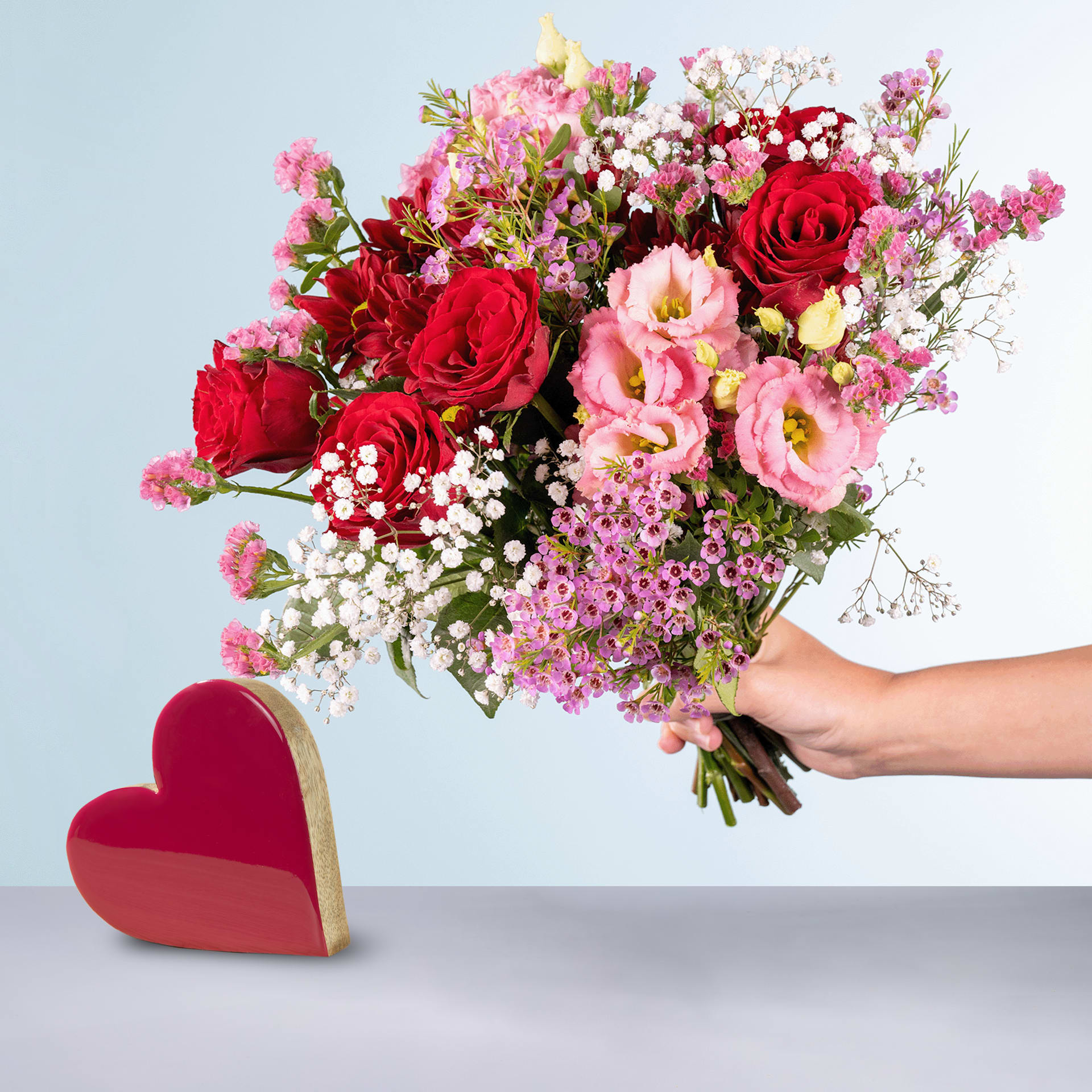 Eine Person hält einen leuchtenden Strauß roter Rosen, rosa Nelken und anderer bunter Blumen in den Händen, im Vordergrund ist auf grauem Hintergrund ein rotes herzförmiges Objekt zu sehen.