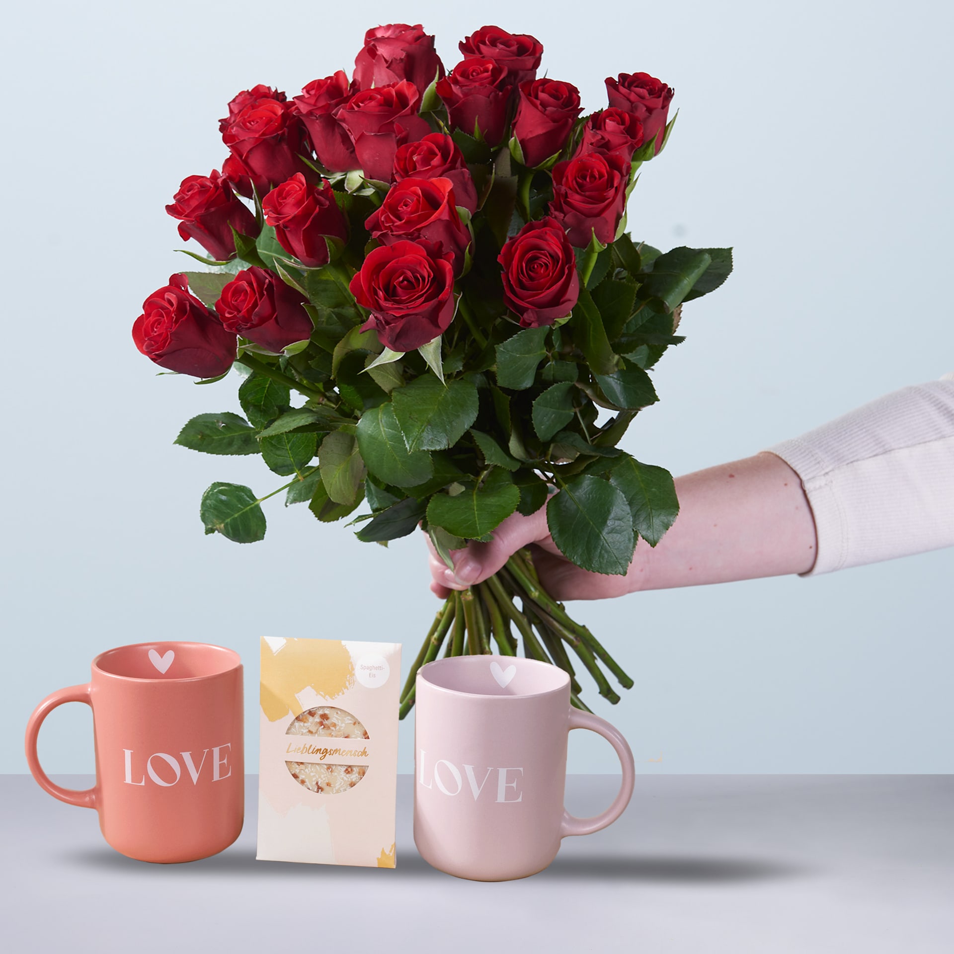 Ein Strauß leuchtend roter Rosen in den Händen einer Person, begleitet von zwei Tassen mit der Aufschrift „LOVE“ auf einem schlichten Hintergrund.