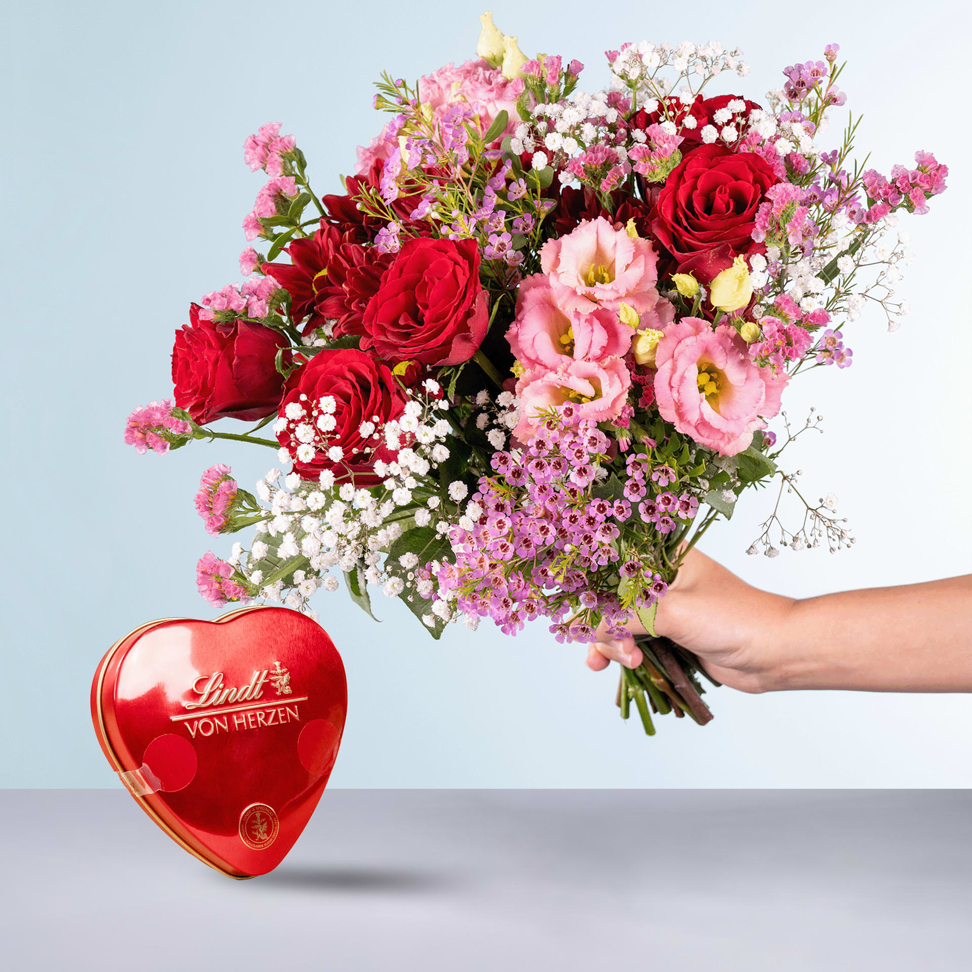 Eine Person hält einen leuchtenden Strauß roter Rosen, rosa Nelken und anderer bunter Blumen in den Händen, im Vordergrund ist vor einem schlichten Hintergrund eine herzförmige Lindt-Schokoladenschachtel zu sehen.