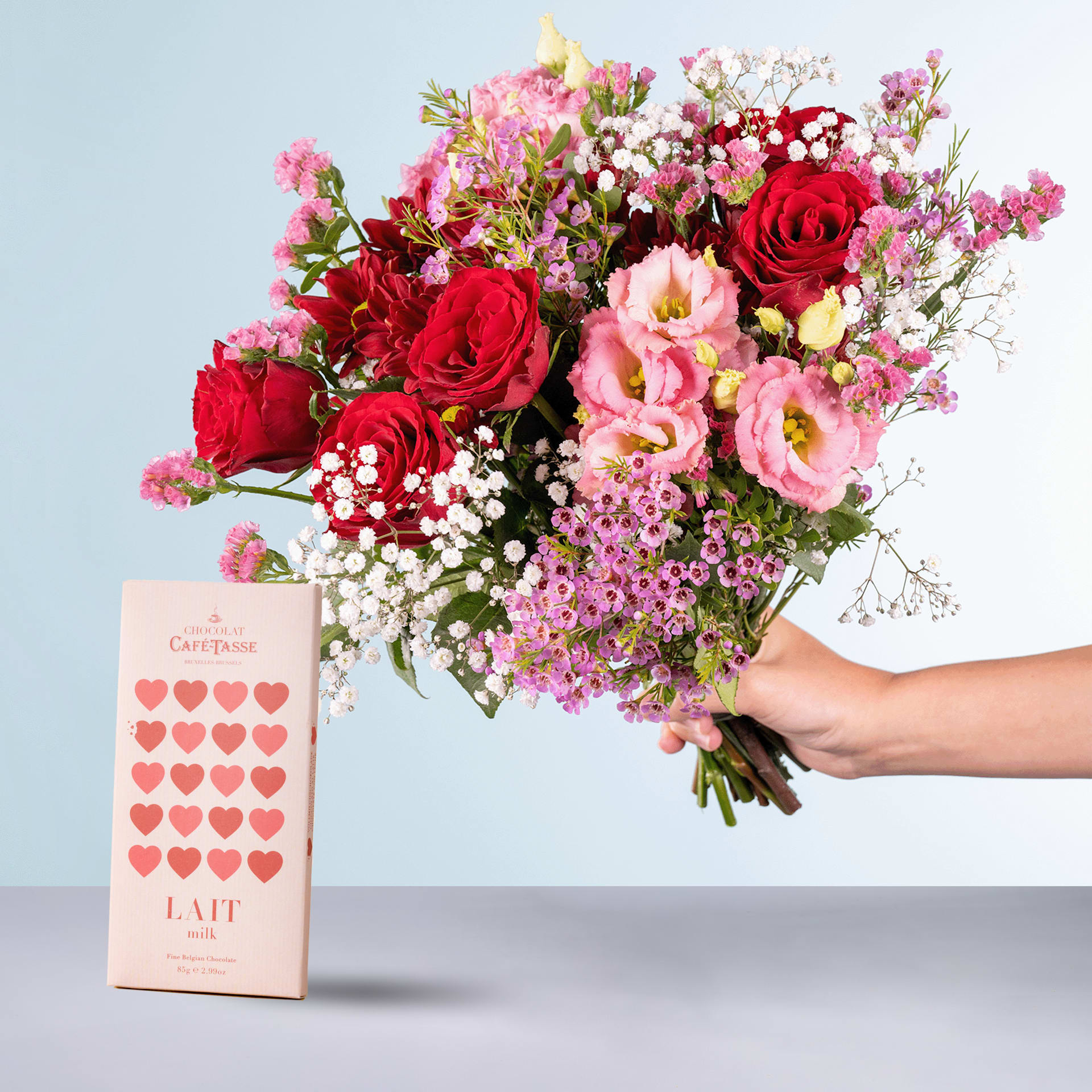 Eine Person hält einen farbenfrohen Strauß roter Rosen, rosa Nelken und anderer bunter Blumen in den Händen, begleitet von einer rosa Geschenkbox mit herzförmigem Muster.