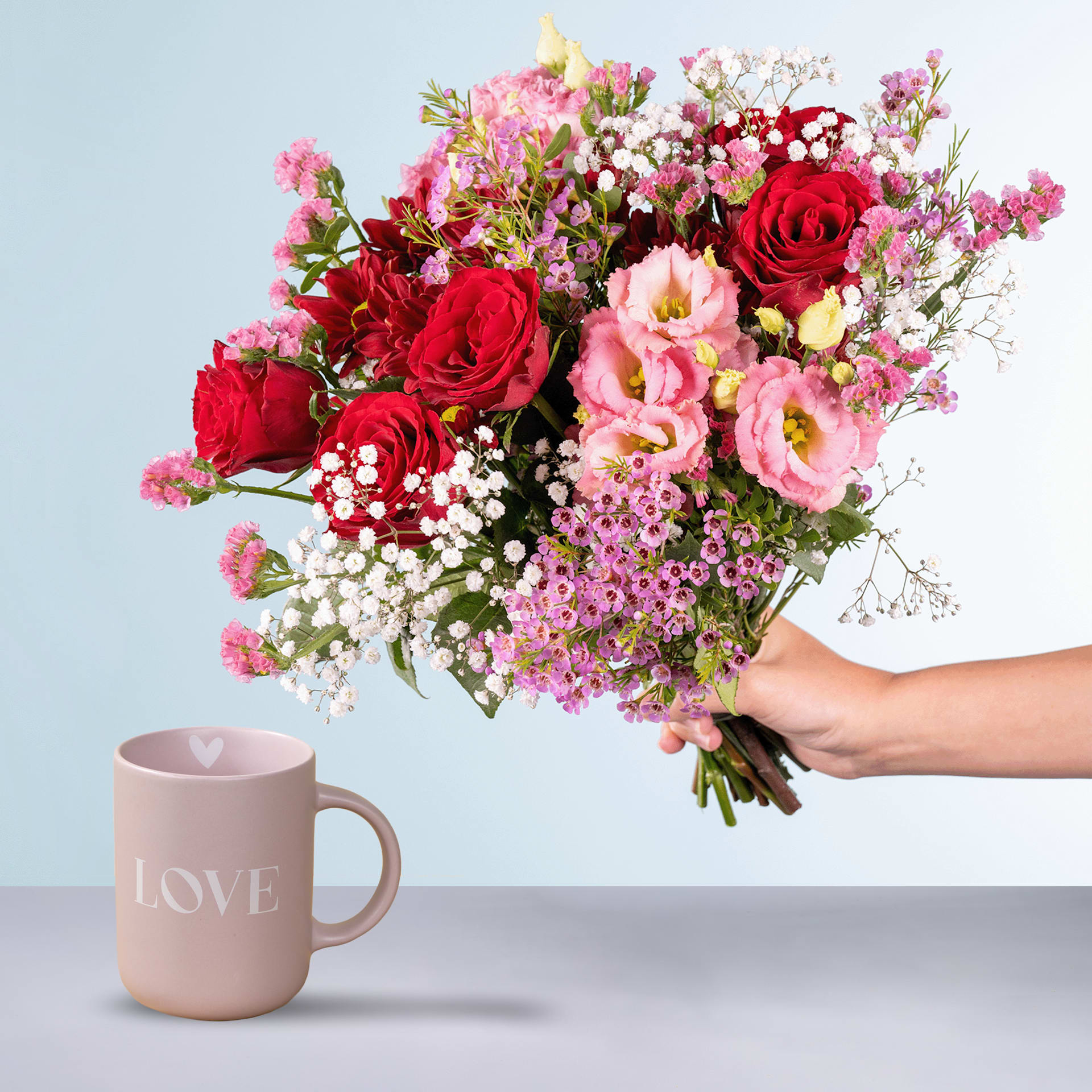 Ein leuchtender Strauß roter Rosen, rosa Nelken und zarter Schleierkrautblüten wird in den Händen einer Person gehalten. Vor einem schlichten hellblauen Hintergrund ist eine rosa Tasse mit dem Wort „LOVE“ im Vordergrund zu sehen.