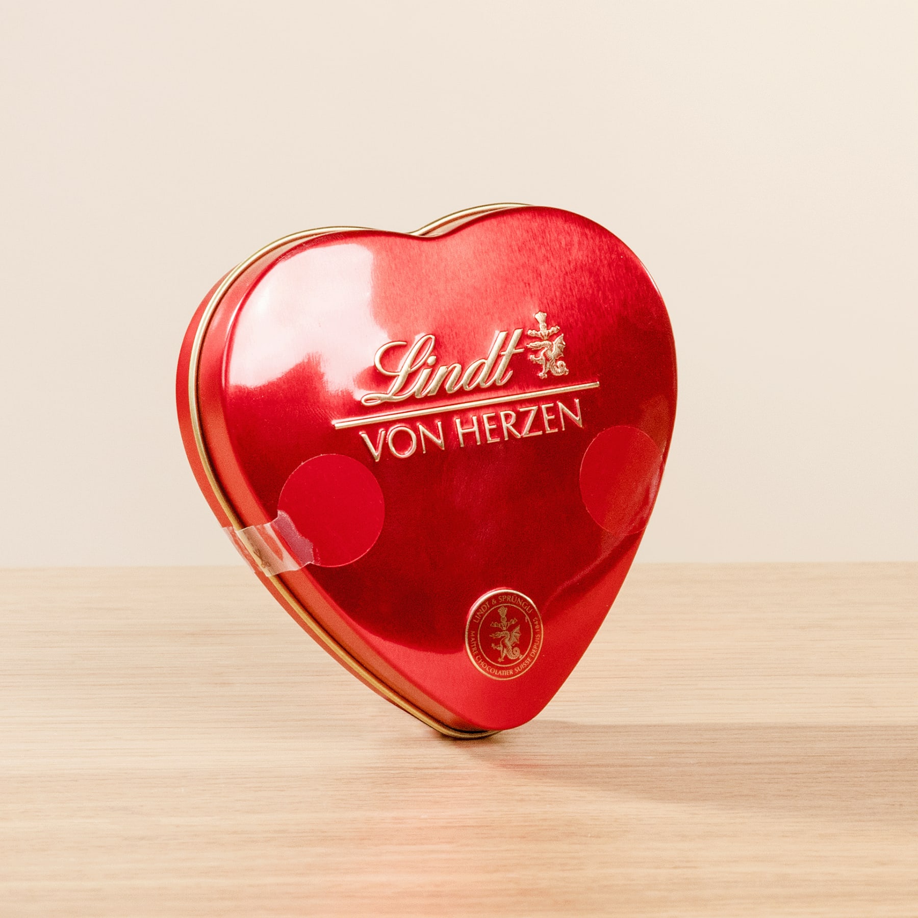 Eine rote herzförmige Dose mit Lindt-Pralinen „Von Herzen“ steht auf einer Holzoberfläche.