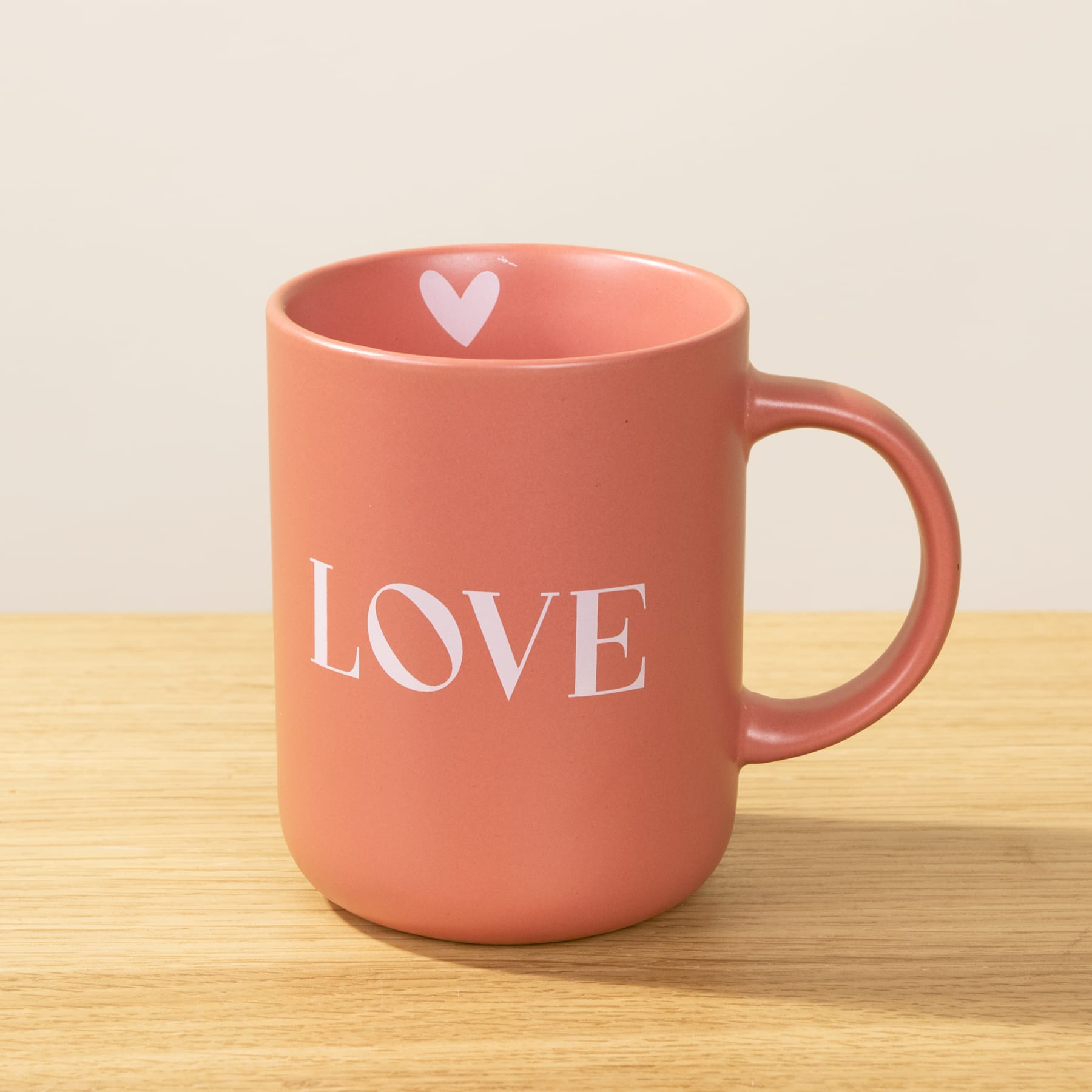 Eine rosa Keramiktasse mit dem Aufdruck „LOVE“ steht auf einer Holzoberfläche vor einem schlichten Hintergrund.