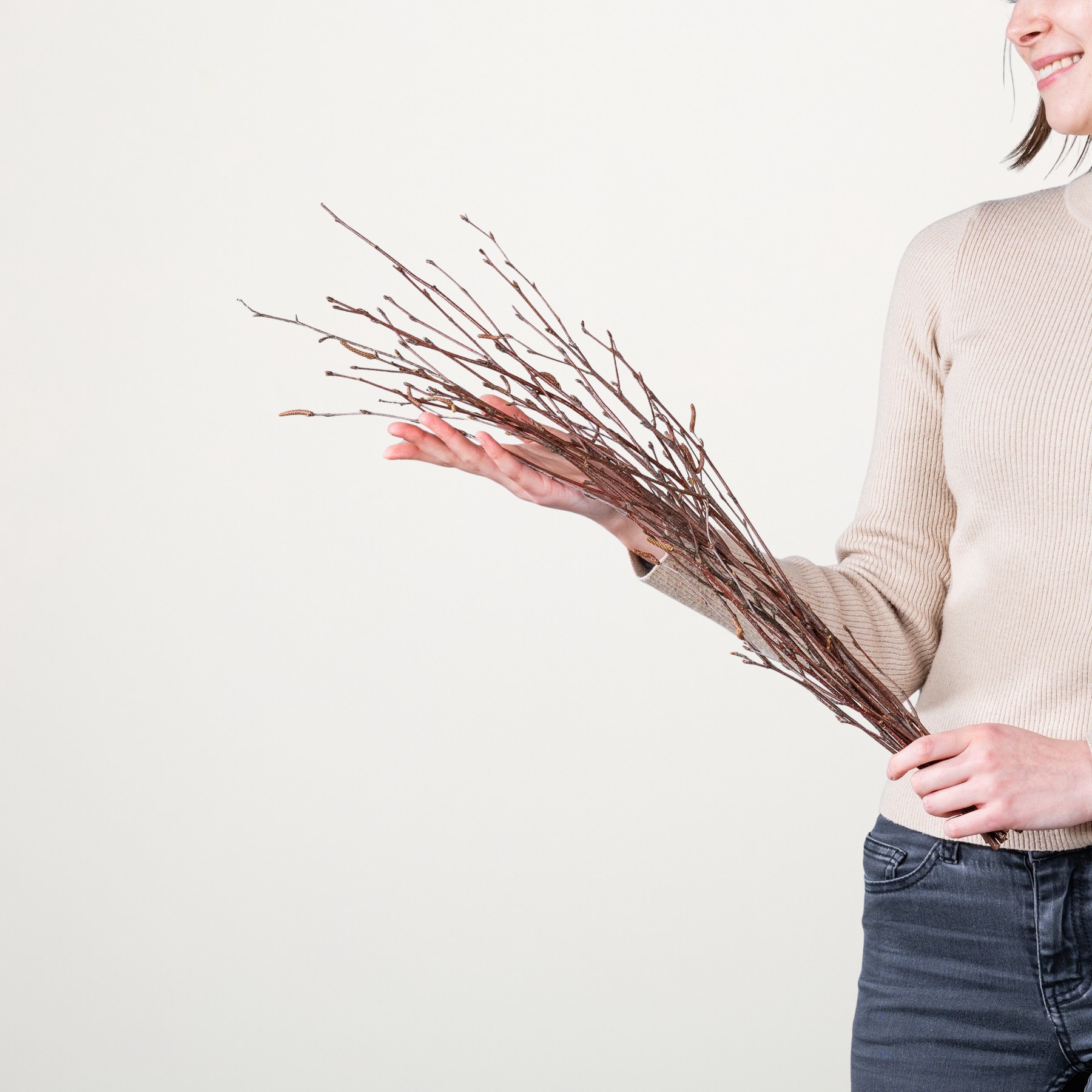 Eine Person in einem beigen Pullover hält ein Bündel kahler Äste vor einem schlichten weißen Hintergrund.