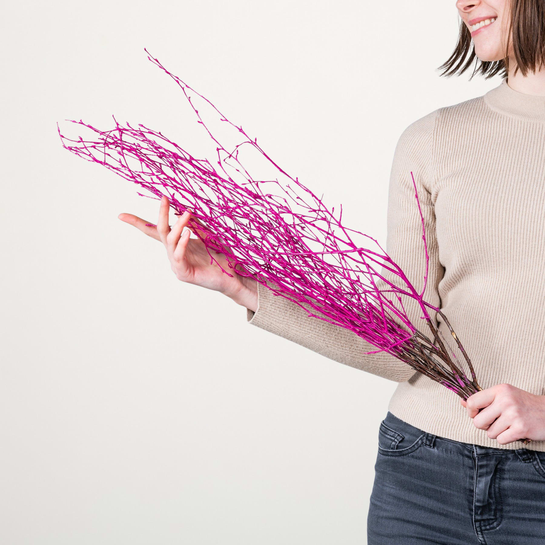Eine Person in einem beigen Pullover hält einen großen Strauß leuchtend rosa Zweige vor einem schlichten weißen Hintergrund.