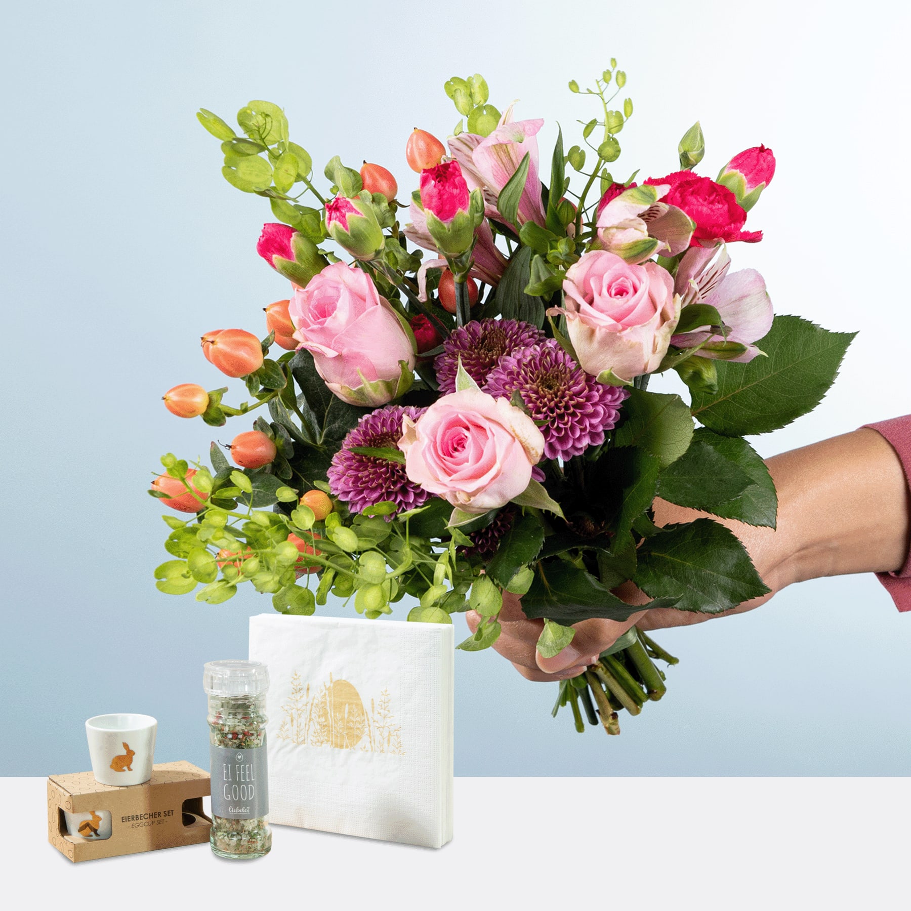 Eine Person hält einen farbenfrohen Strauß rosa Rosen, lila Chrysanthemen und Grünpflanzen in den Händen, begleitet von einer Geschenkbox und einer dekorativen Glasvase auf hellblauem Hintergrund.