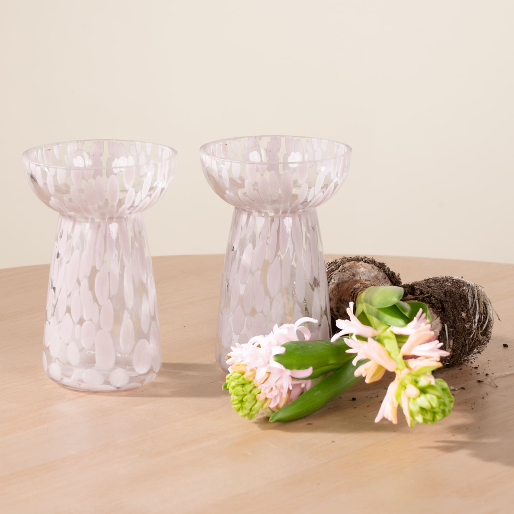 Zwei klare Glasvasen mit strukturiertem Muster stehen auf einer Holzoberfläche, begleitet von einer kleinen nestartigen Struktur und einem Strauß leuchtend grüner und weißer Blumen.