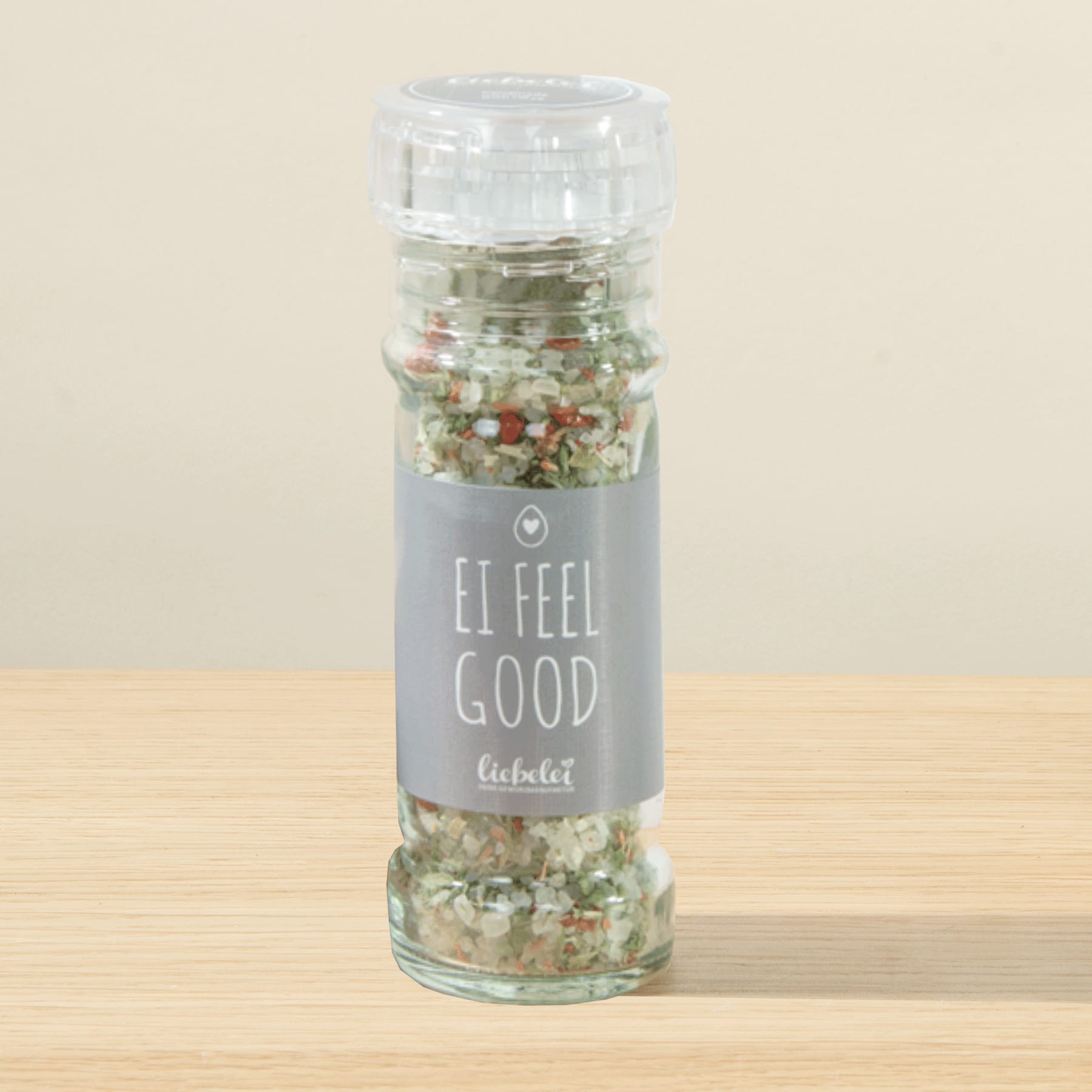 Eine Gewürzmühle aus Glas, gefüllt mit einer bunten Gewürzmischung, steht auf einer Holzoberfläche vor einem hellen Hintergrund.