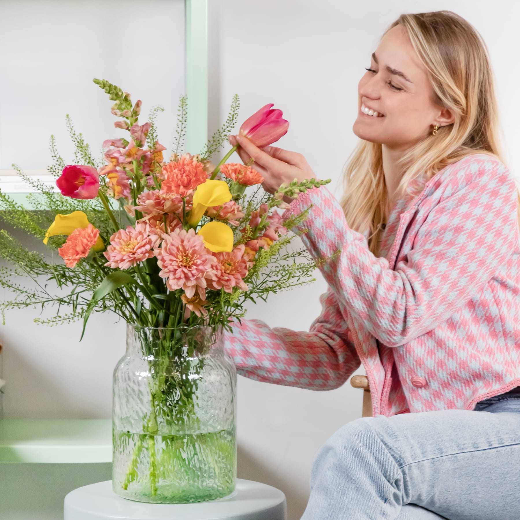 Eine lächelnde junge Frau in einem rosa-weiß karierten Pullover arrangiert einen farbenfrohen Strauß bunter Blumen in einer Glasvase vor einem schlichten weißen Hintergrund.