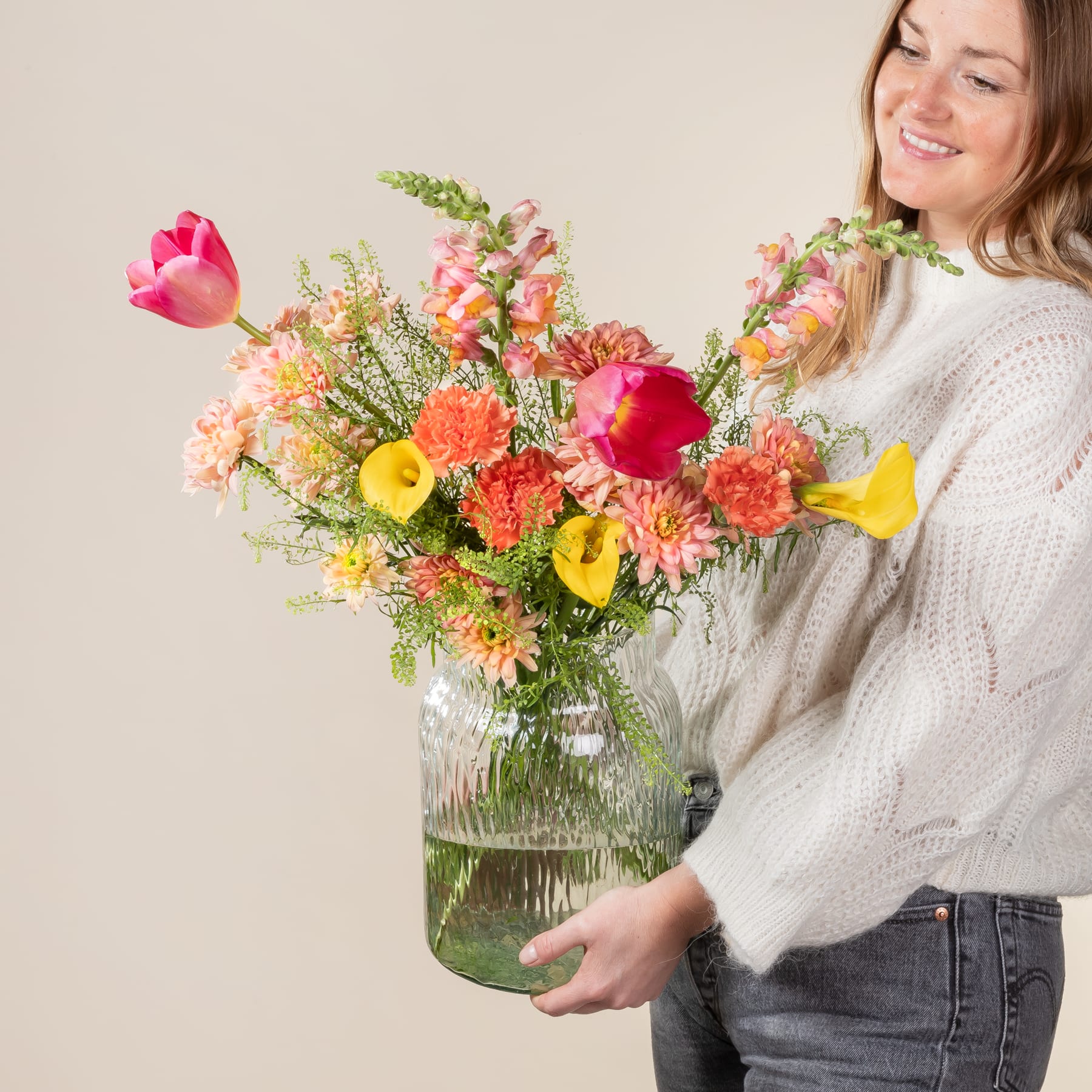 Eine lächelnde Frau hält vor einem neutralen Hintergrund einen großen Strauß leuchtend bunter Blumen in einer Glasvase.