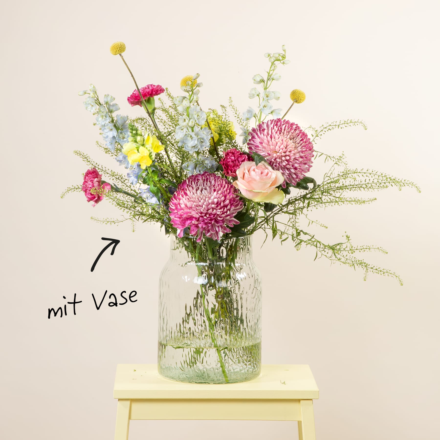 Ein Strauß leuchtend rosa und gelber Blumen, darunter Chrysanthemen und Rosen, wird in einer Glasvase auf einem gelben Holzhocker vor einem schlichten weißen Hintergrund präsentiert.