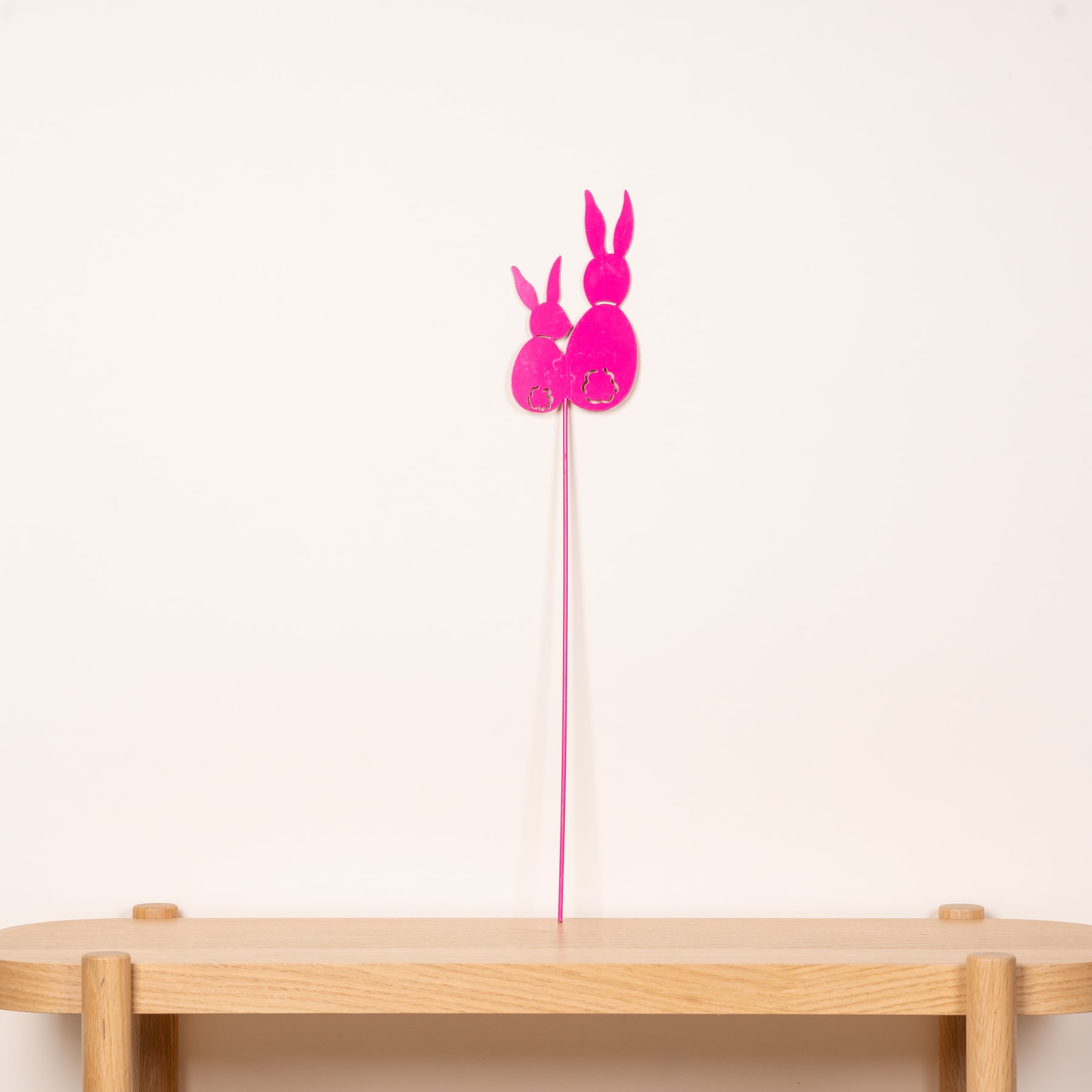 Auf einem Holztisch vor einem schlichten Hintergrund steht eine leuchtend rosa Dekoration in Hasenform.