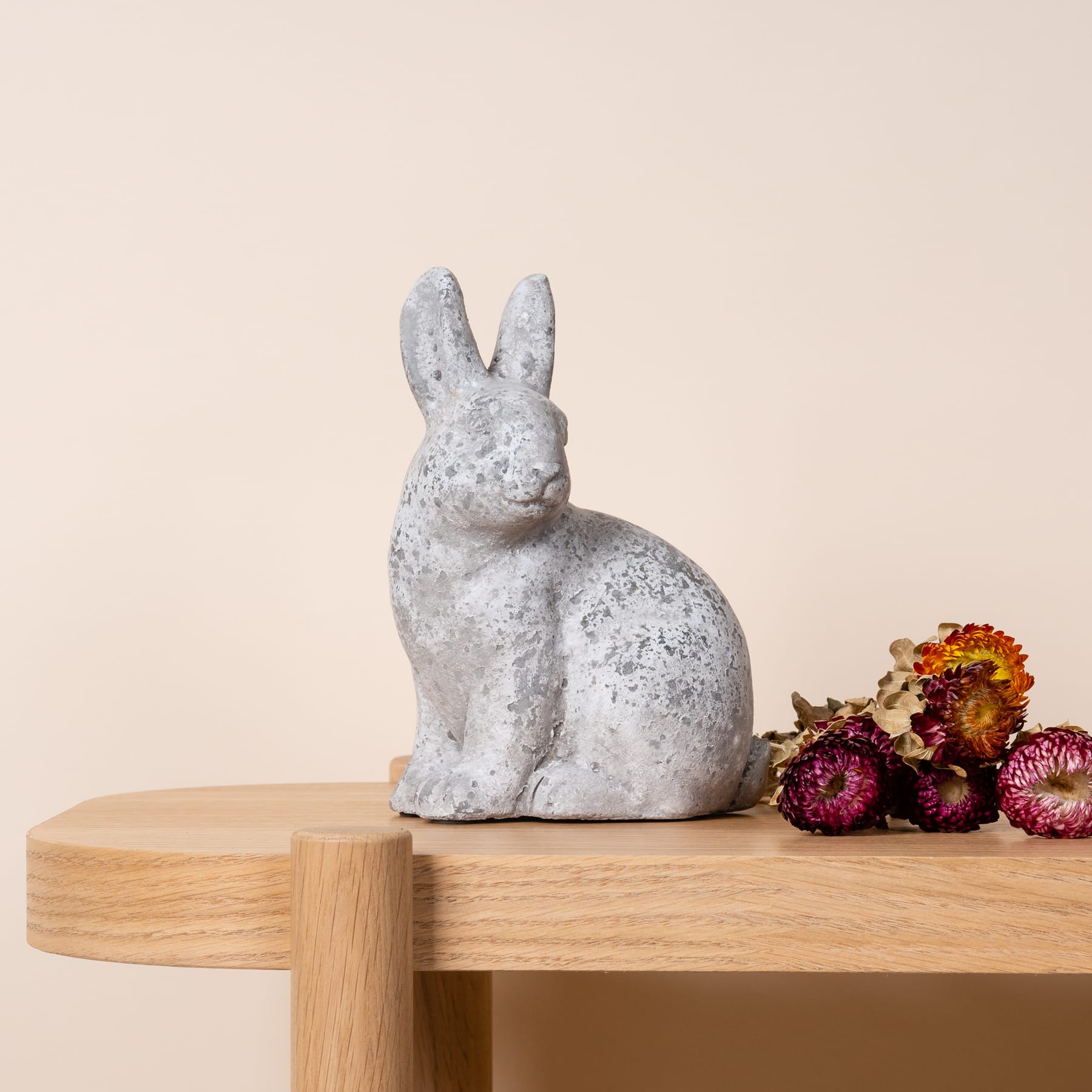Eine Kaninchenfigur aus Beton sitzt auf einer Holzoberfläche, umgeben von leuchtend rosa und violetten Blumen.
