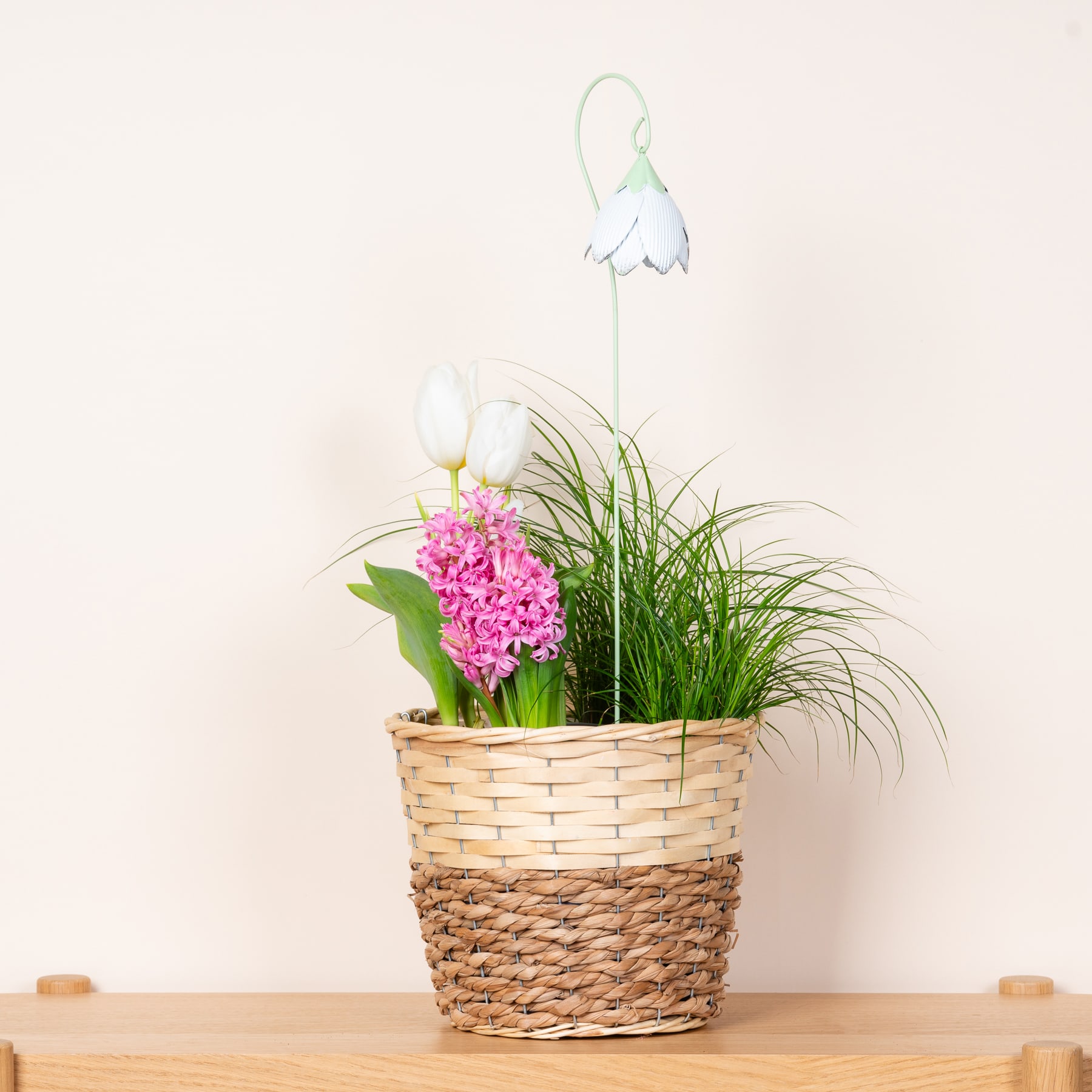 Ein geflochtener Korb voller leuchtend rosa Blumen, üppigem grünem Laub und einer zarten weißen Blume steht auf einer Holzoberfläche vor einem schlichten Hintergrund.