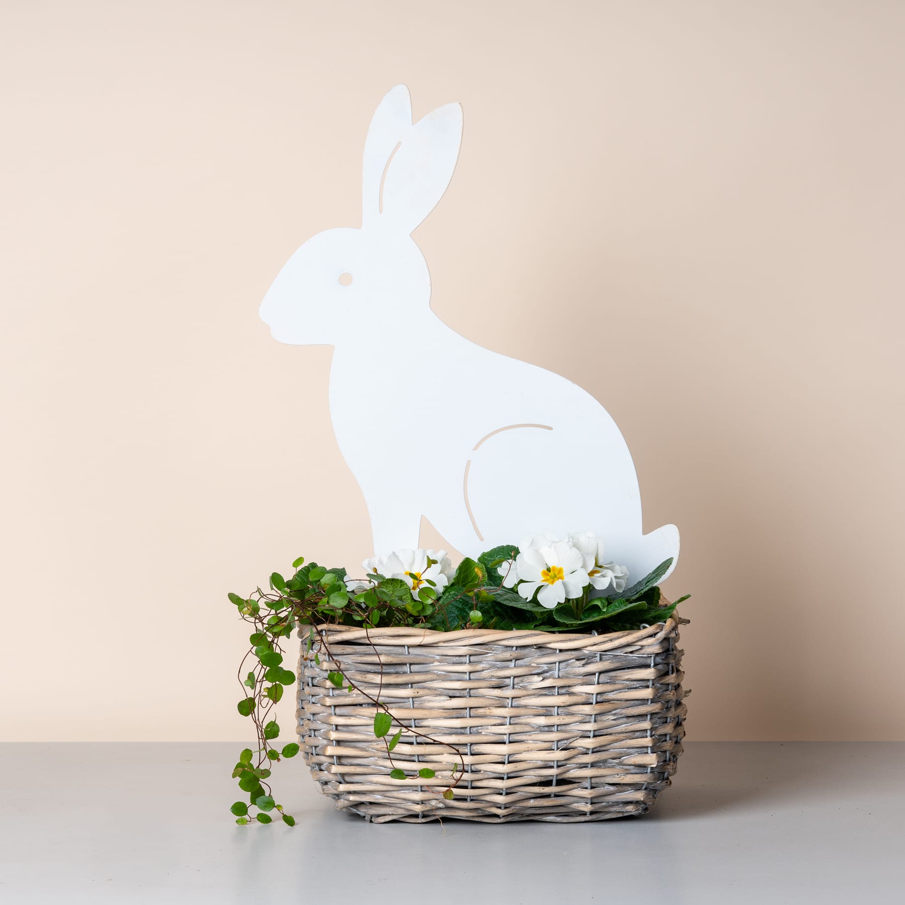 Die weiße Silhouette eines Kaninchens sitzt auf einem Weidenkorb voller üppigem Grün und zarten weißen Blumen vor einem neutralen beigen Hintergrund.