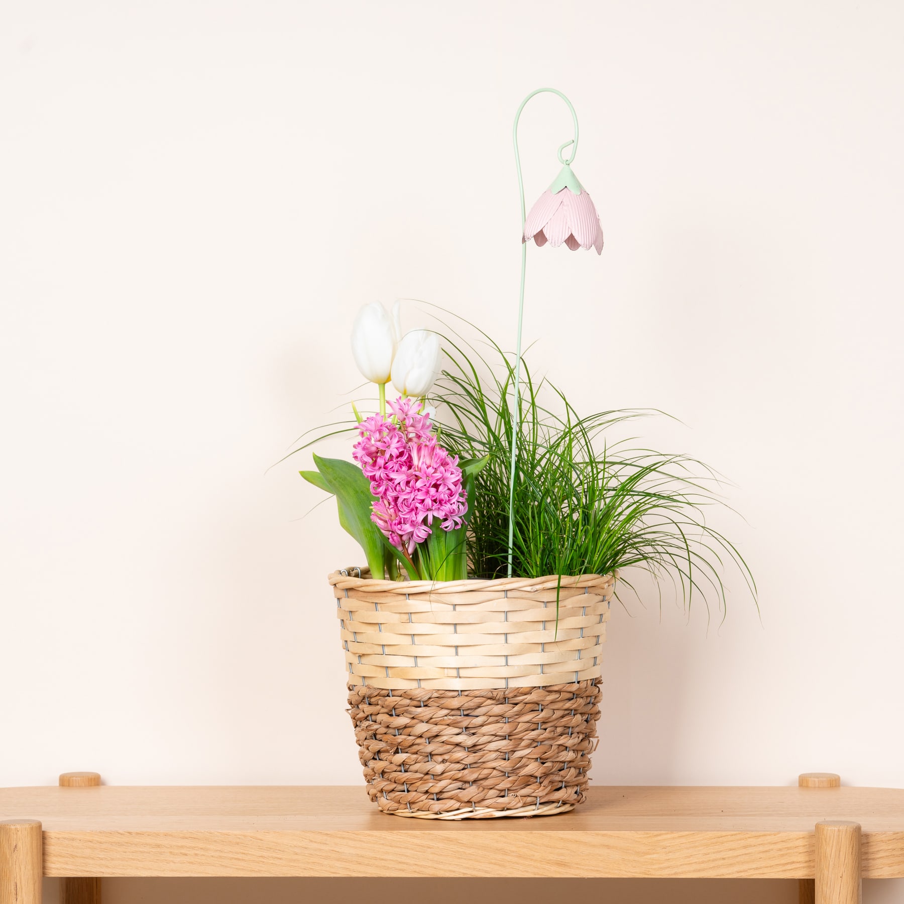 Ein geflochtener Korb voller leuchtend rosa Blumen, üppigem grünem Laub und einer zarten weißen Blume hängt vor einem schlichten weißen Hintergrund.