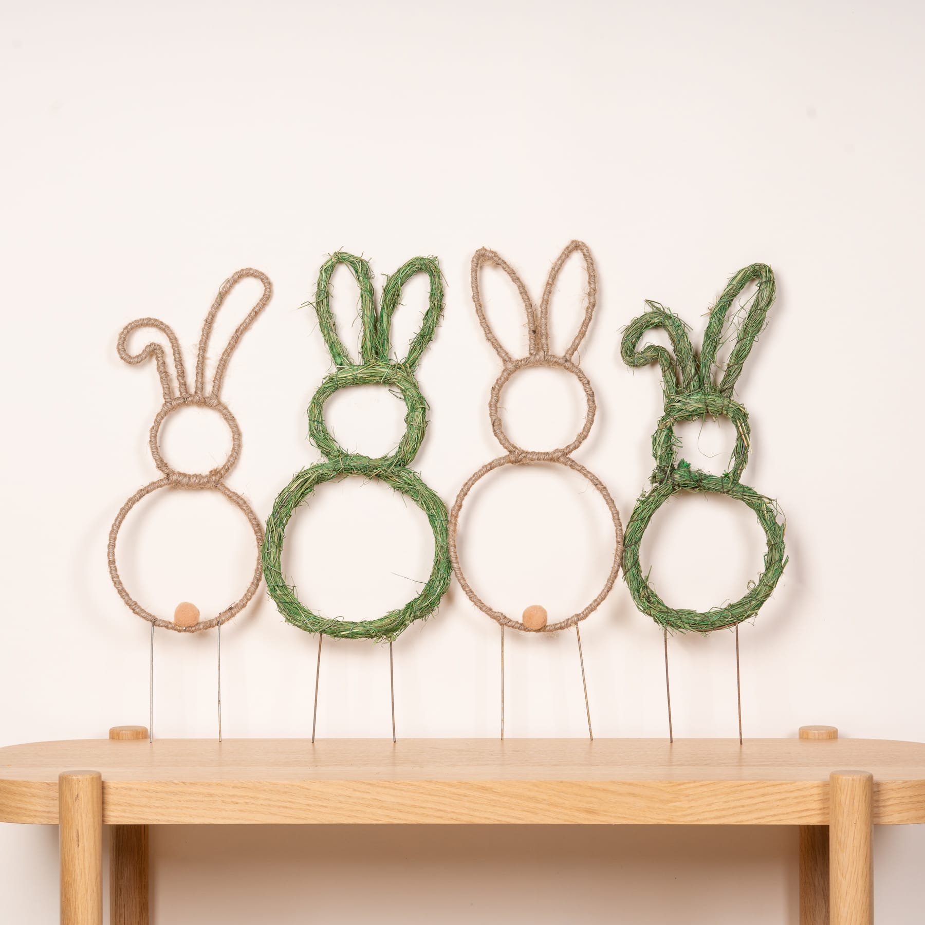Gehäkelte Hasenohren in verschiedenen Grün- und Beigetönen sind auf einem Holztisch vor einem schlichten Hintergrund ausgestellt.