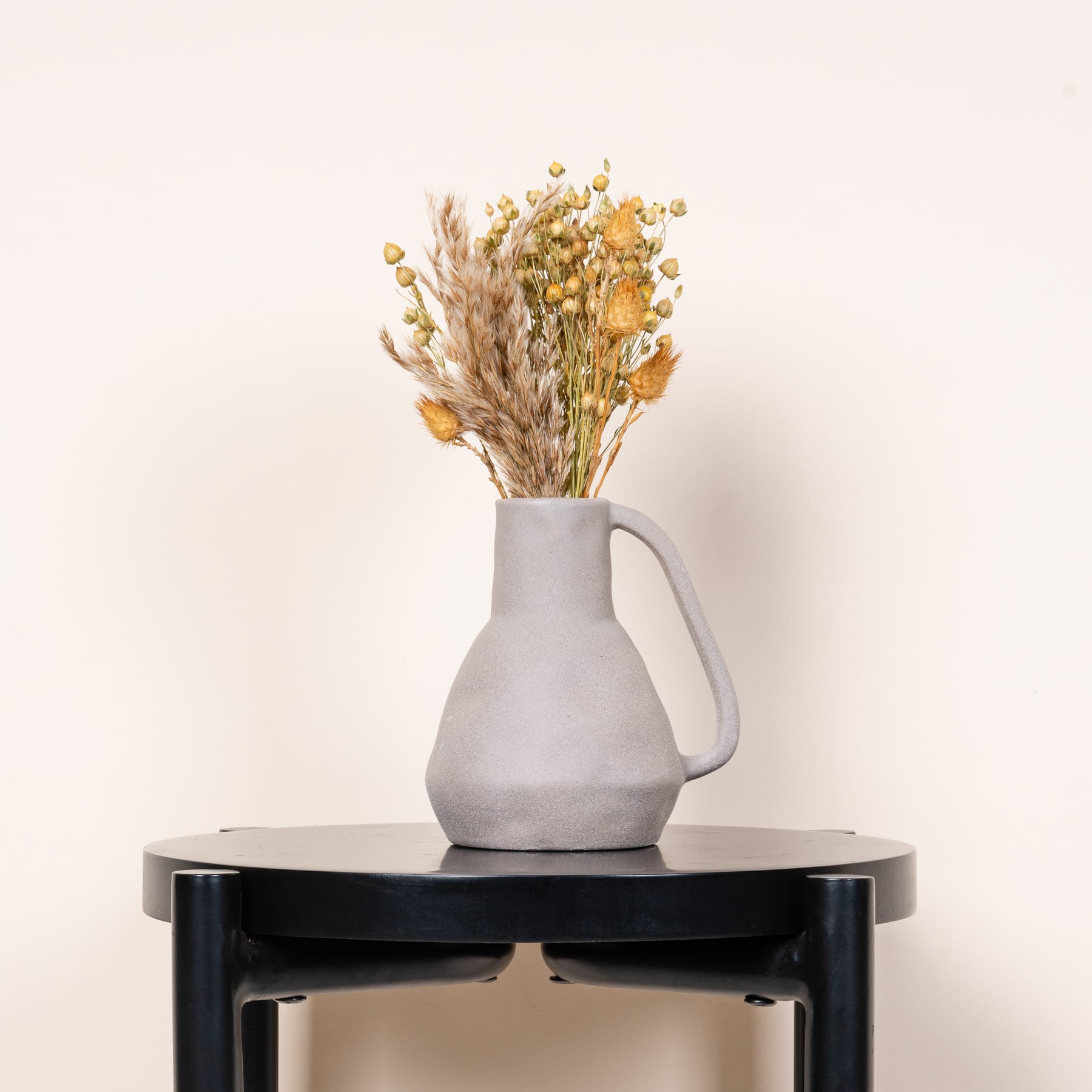 Ein einfaches, minimalistisches Arrangement aus einem grauen Keramikkrug, gefüllt mit einem Strauß getrockneter Blumen und Gräser, vor einem schlichten, hellen Hintergrund.