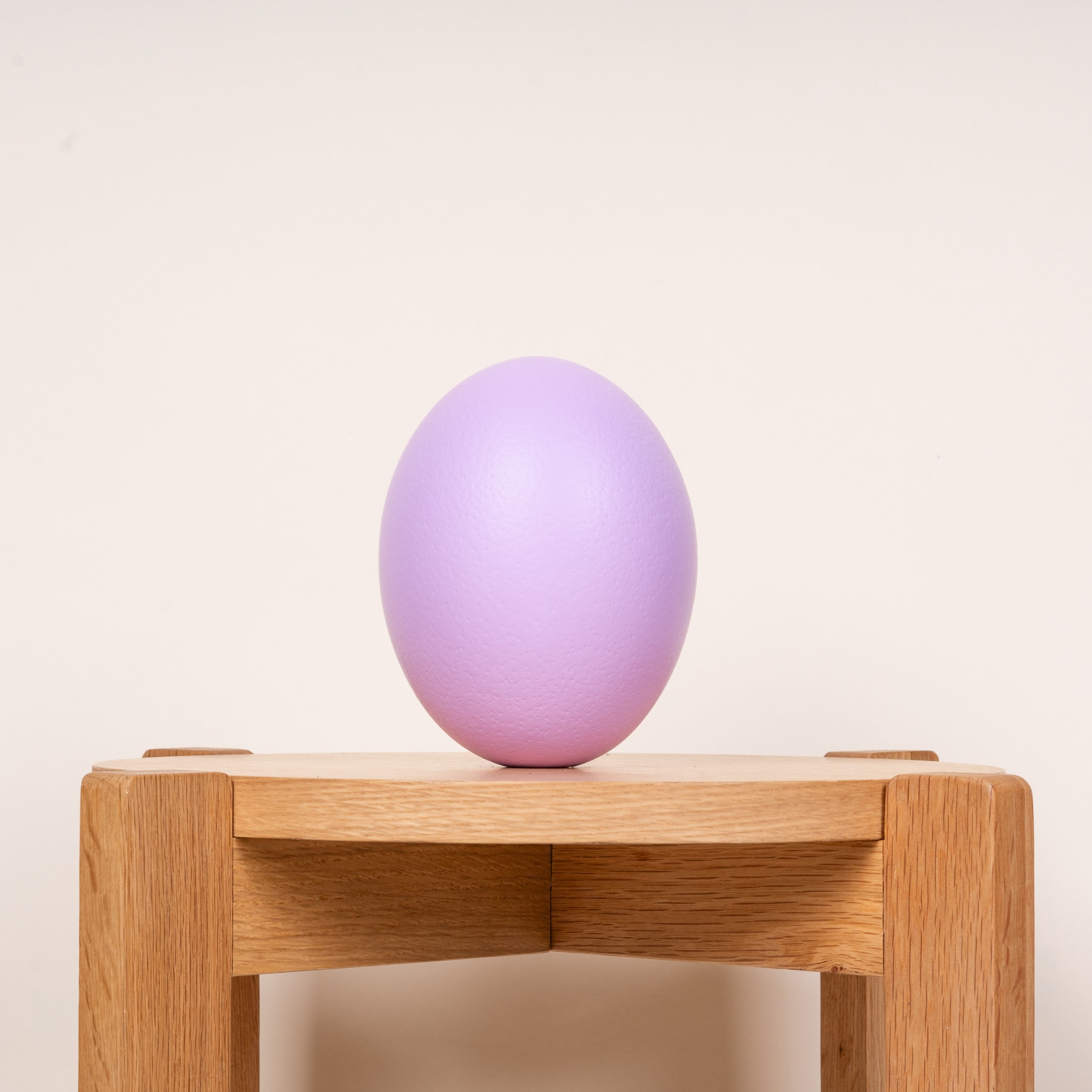 Ein pastellviolettes, eiförmiges Objekt ruht auf einem Holzständer vor einem schlichten, hellen Hintergrund.