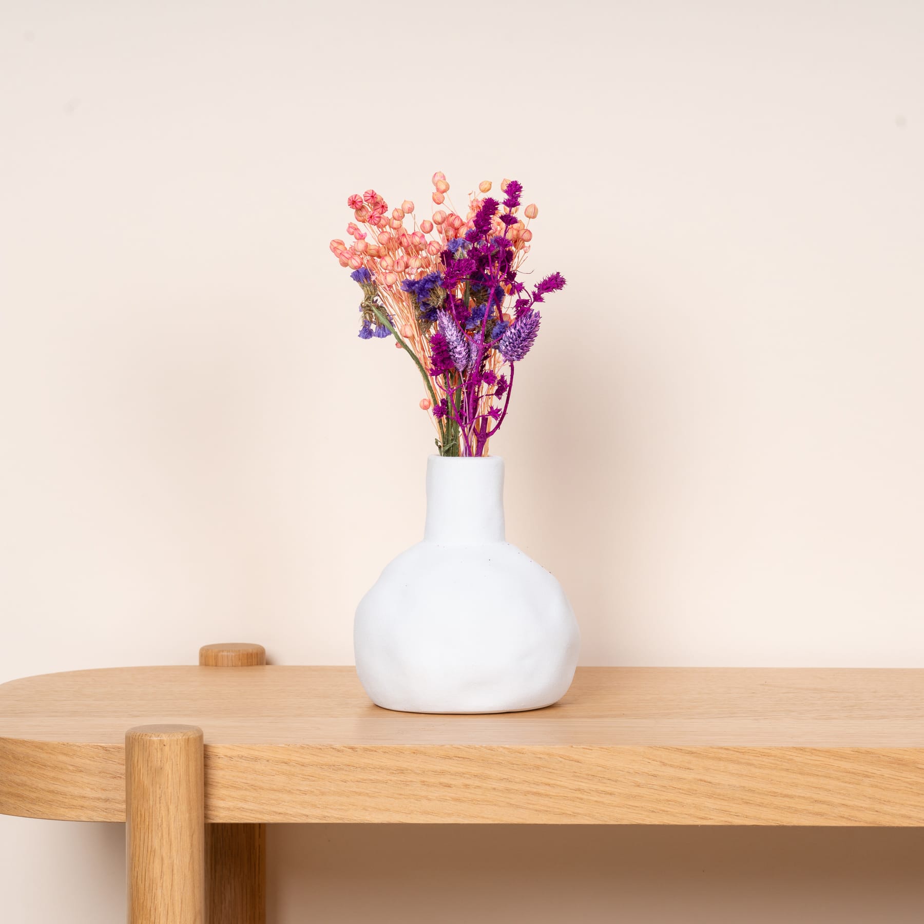 Eine weiße Keramikvase mit einem farbenfrohen Strauß getrockneter Blumen steht auf einem Holzregal vor einer schlichten, hellen Wand.