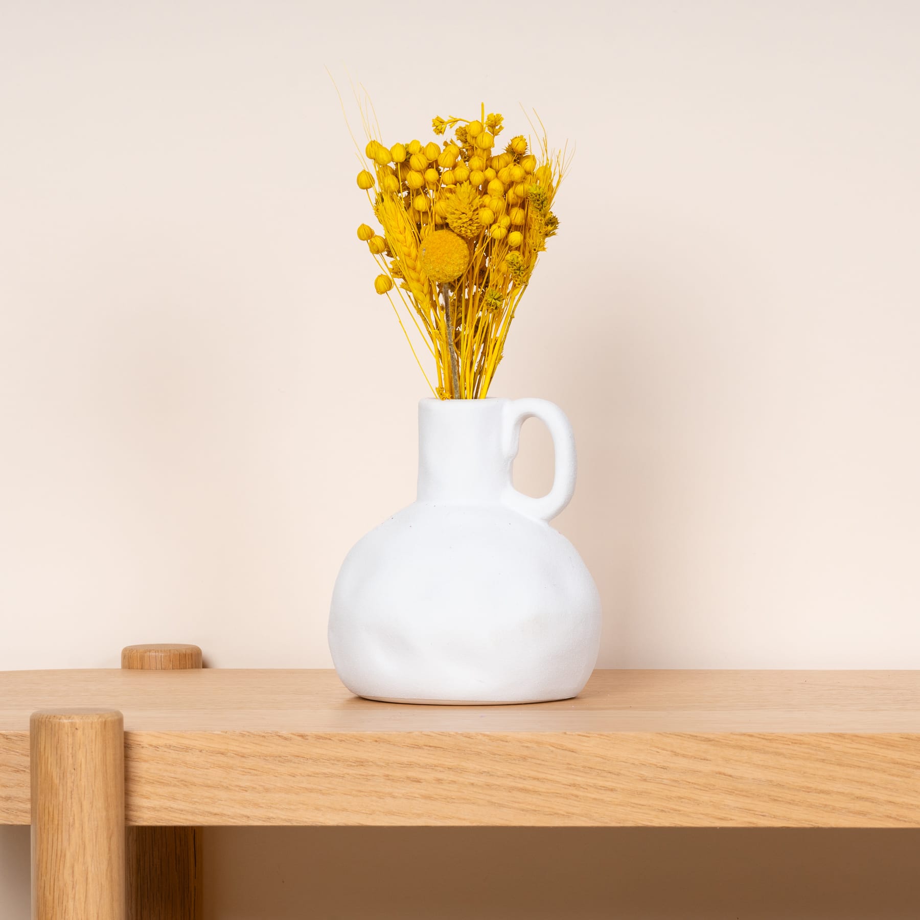 Eine weiße Keramikvase mit leuchtend gelben Blumen steht auf einem Holztisch vor einem schlichten beigen Hintergrund.