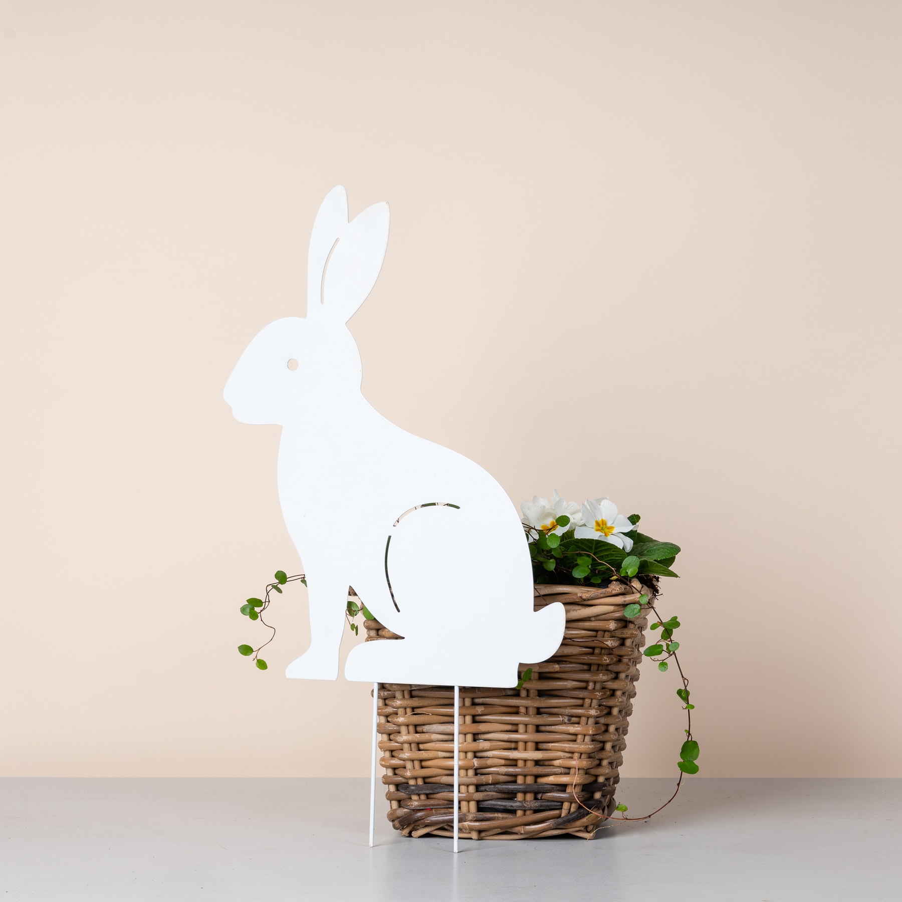 Die Silhouette eines weißen Kaninchens aus Holz sitzt in einem geflochtenen Korb voller Grünpflanzen und kleiner weißer Blumen vor einem beigen Hintergrund.