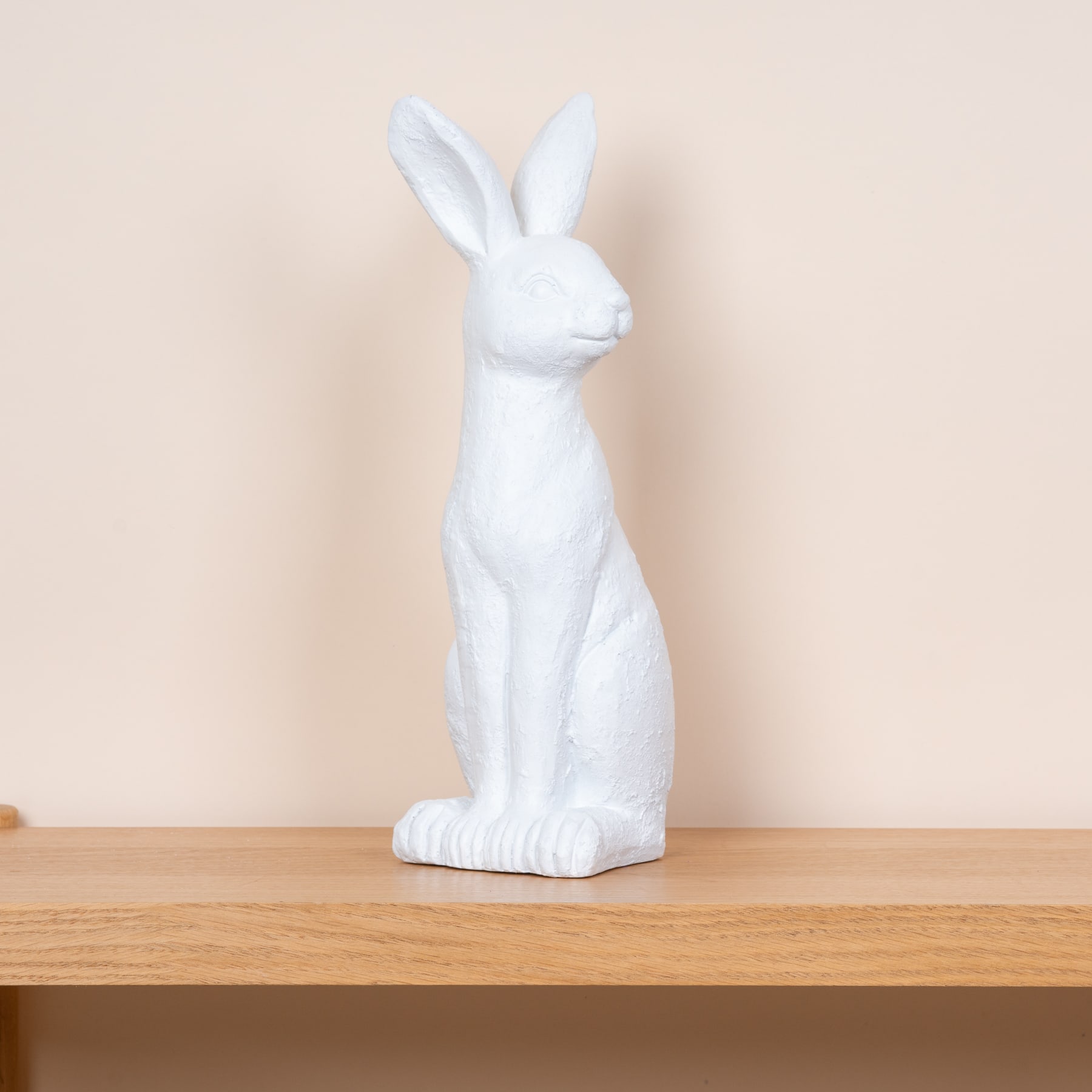 Eine weiße Kaninchenfigur aus Keramik steht auf einem Holzregal vor einer beigen Wand.