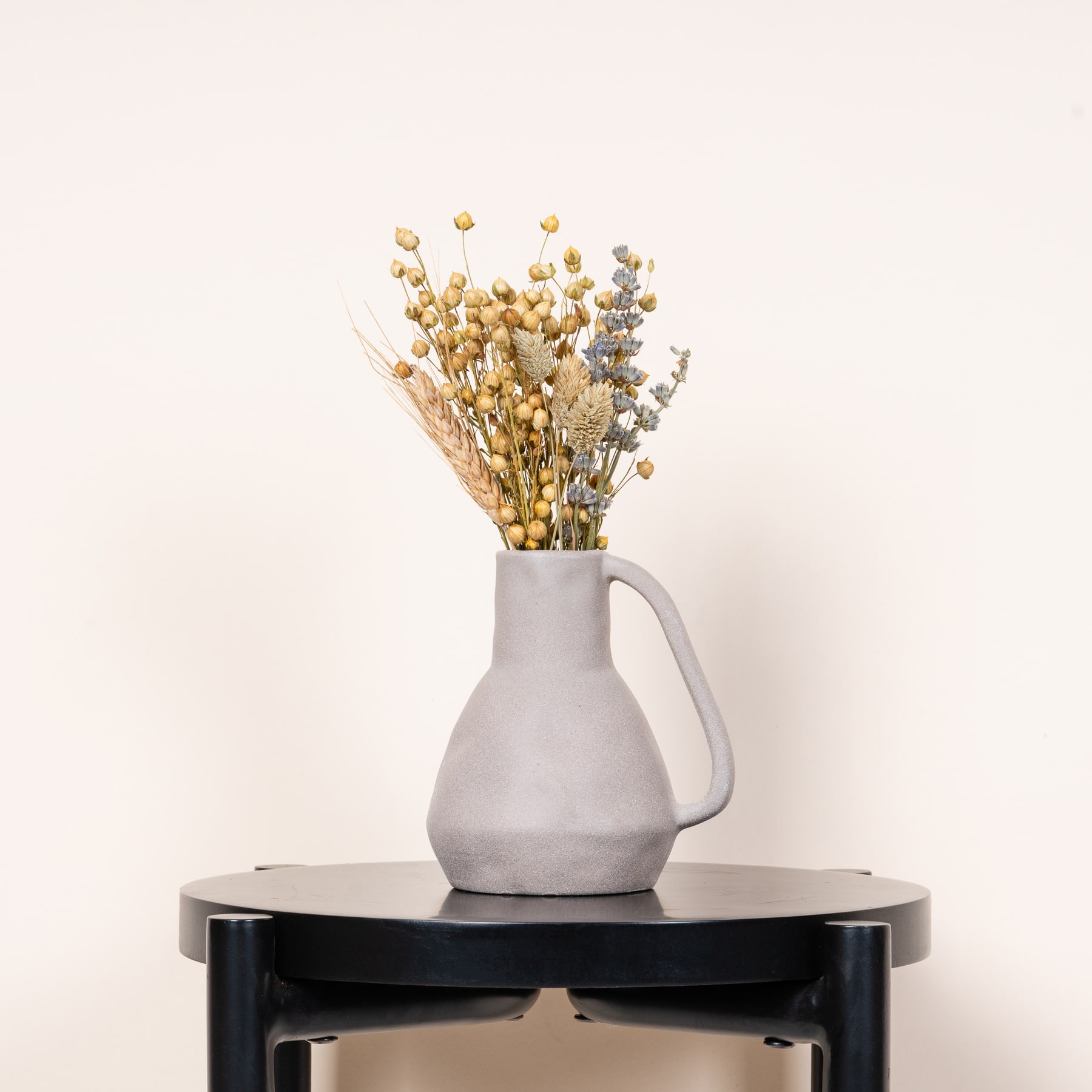 Eine graue Keramikvase mit einem Strauß getrockneter gelber und weißer Blumen steht auf einem dunklen Holztisch vor einem schlichten hellen Hintergrund.