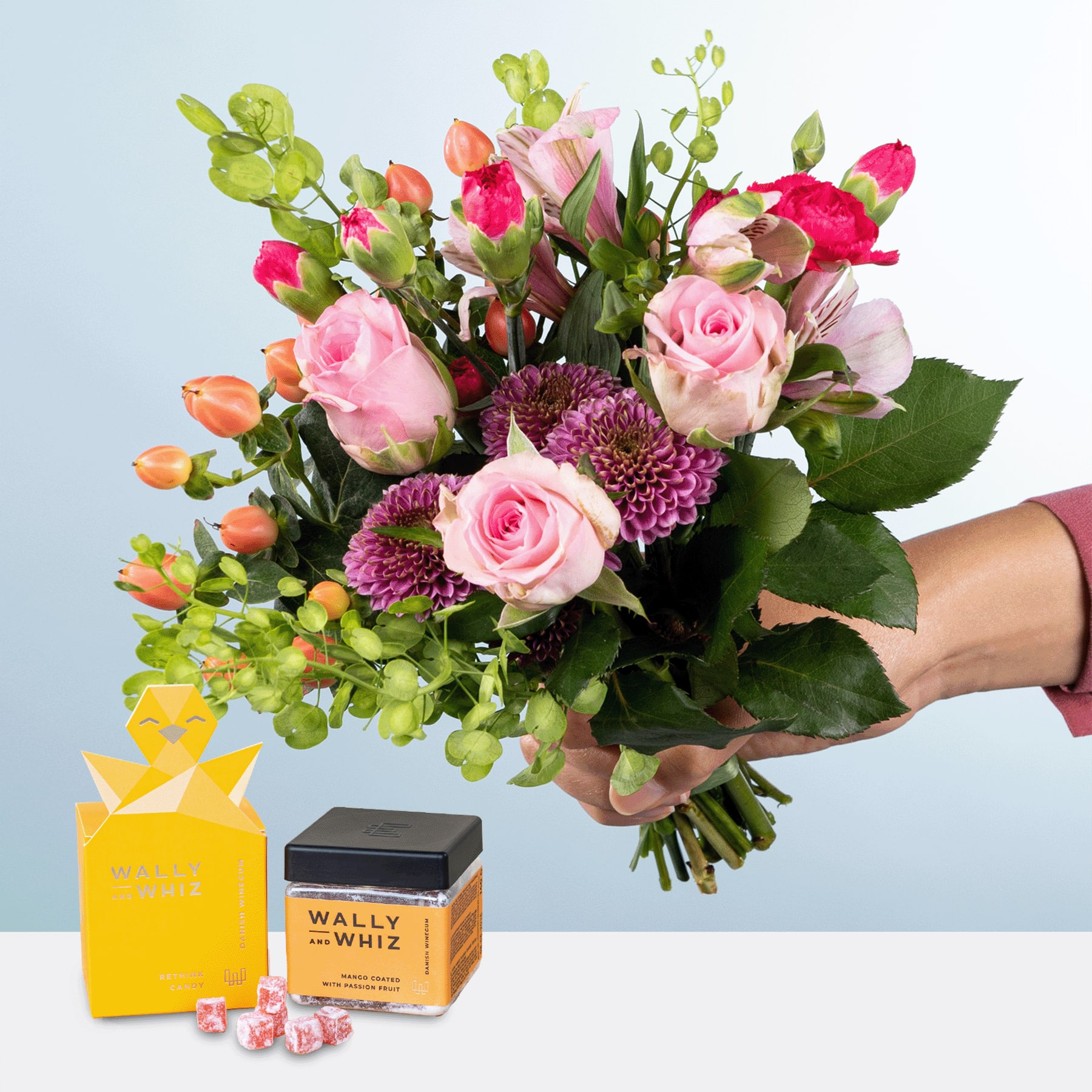 Ein leuchtender Strauß rosa Rosen, lila Chrysanthemen und anderer bunter Blumen in den Händen einer Person, begleitet von einer gelben Geschenkbox und einer Schachtel Wally Whitz-Pralinen auf hellblauem Hintergrund.