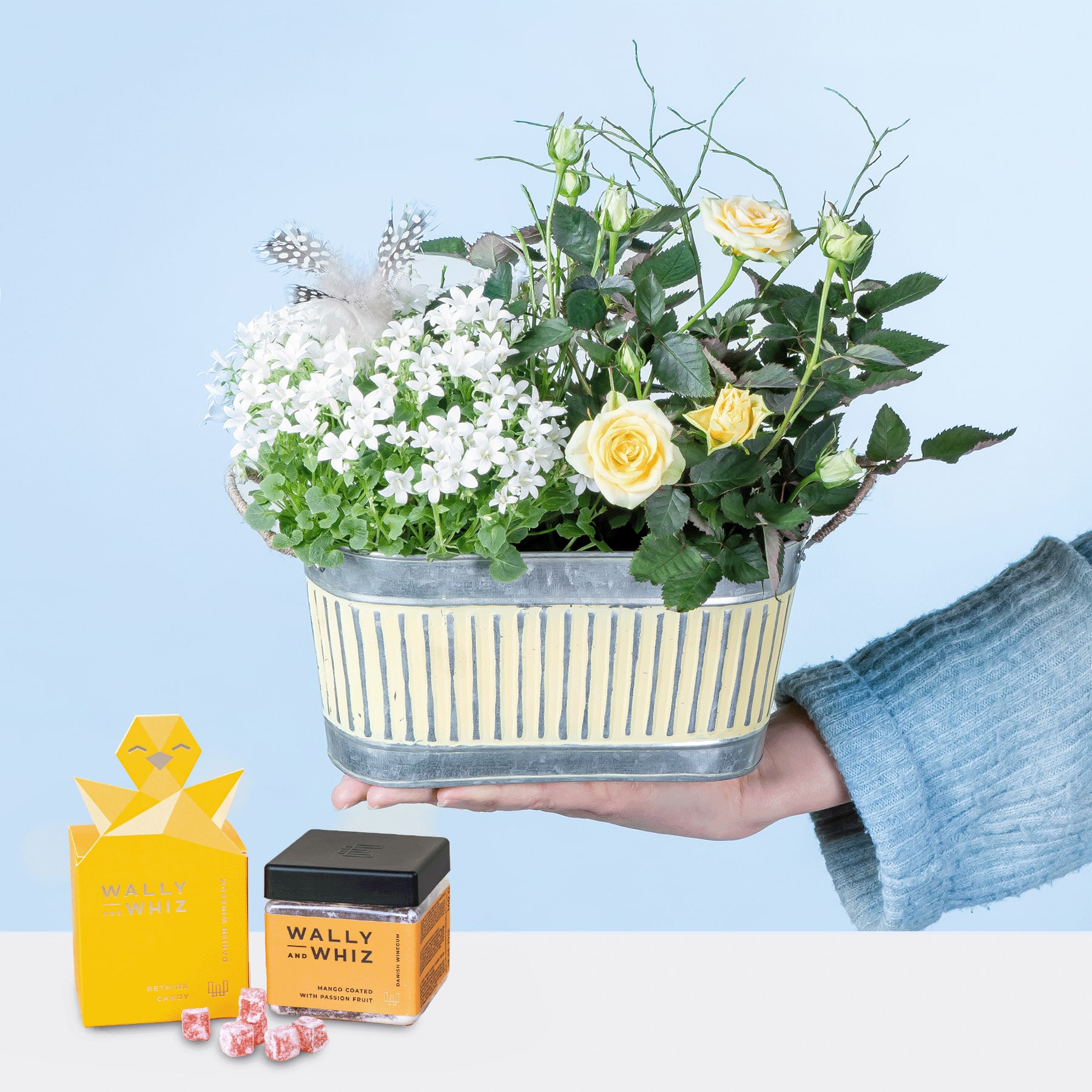 Ein bunter Blumenstrauß mit weißen und gelben Blüten steht in einem gestreiften Blumentopf vor einem schlichten blauen Hintergrund. Im Vordergrund sind eine kleine gelbe Schachtel und einige Süßigkeiten zu sehen.