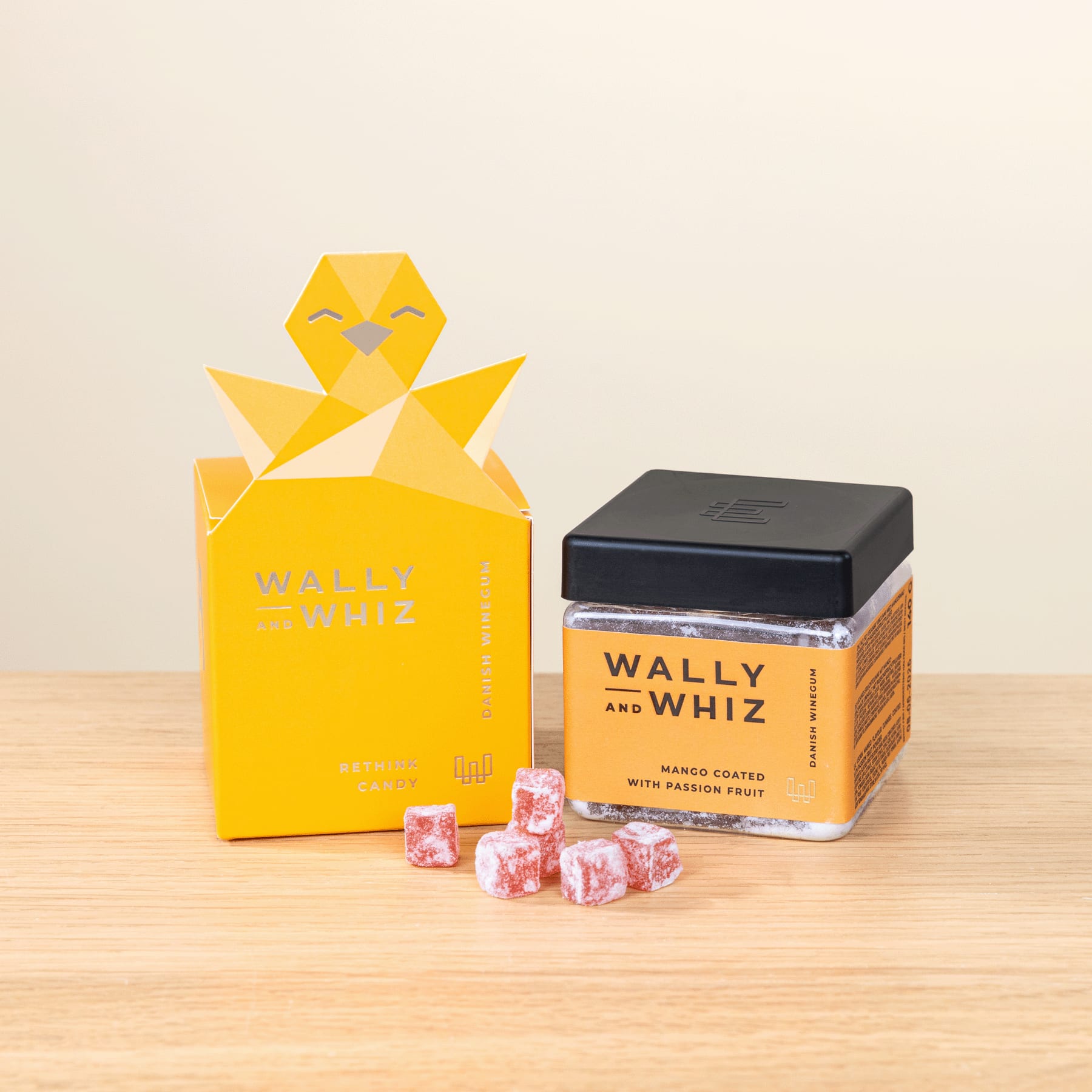 Das Bild zeigt eine gelbe Kükenfigur im Origami-Stil, einen Blechbehälter mit der Aufschrift „Wally and Whiz“ und mehrere rosa Bonbons auf einer Holzoberfläche.