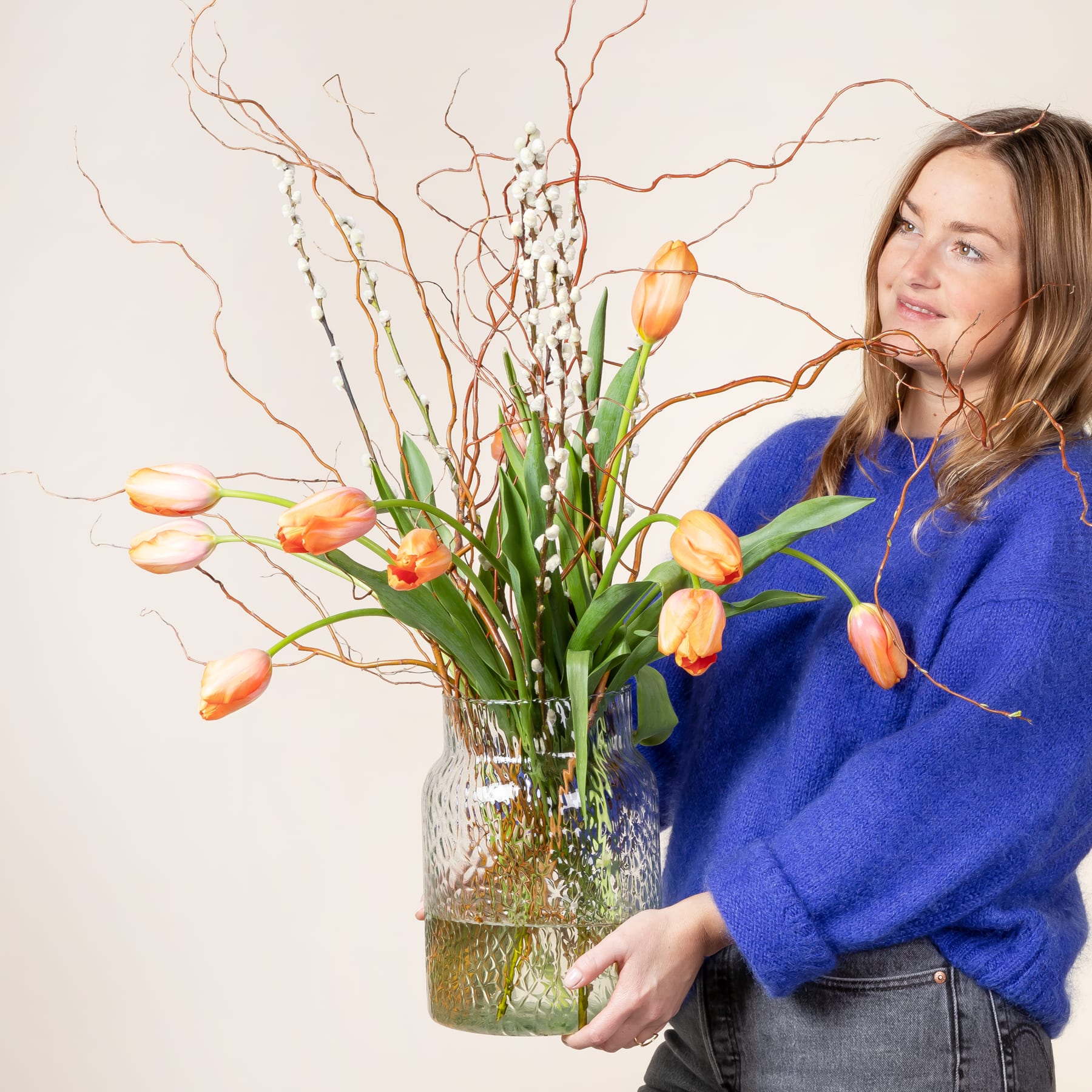 Eine Frau in einem blauen Pullover hält vor einem schlichten weißen Hintergrund eine Vase mit leuchtend orangefarbenen Tulpen und Zweigen.