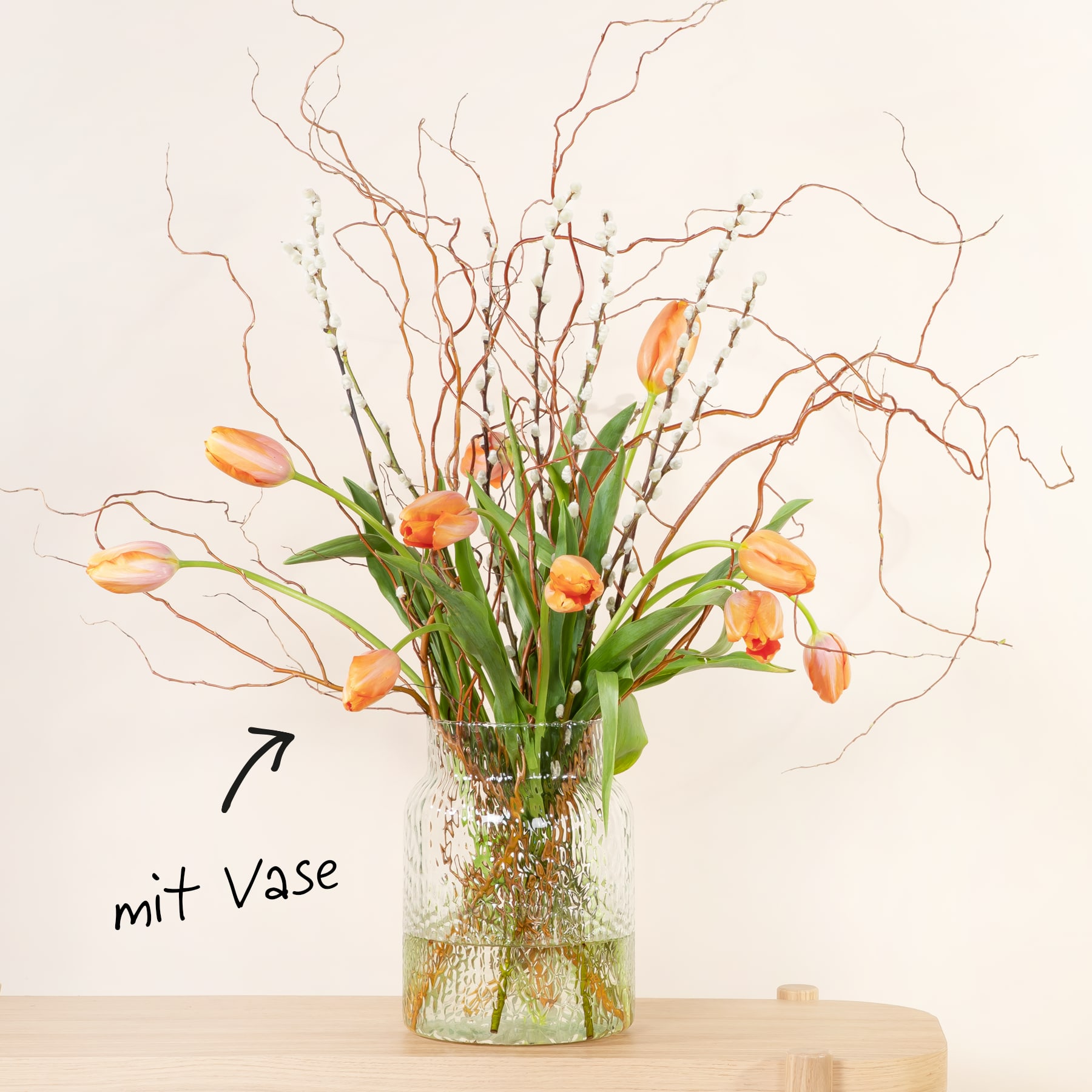 Eine Vase mit leuchtend orangefarbenen Tulpen und Zweigen steht auf einer Holzoberfläche vor einem schlichten weißen Hintergrund.