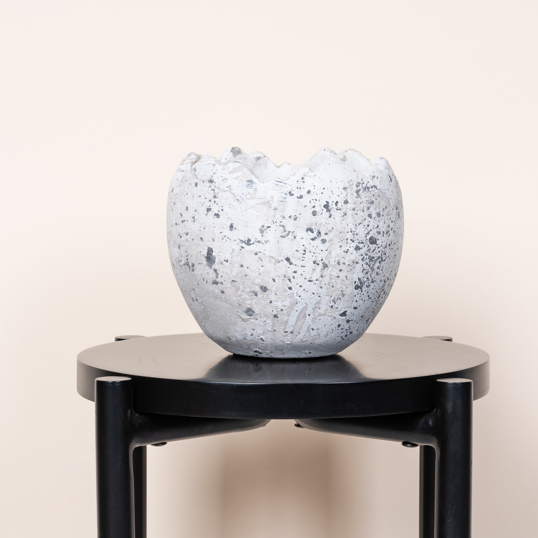 Eine weiße, gesprenkelte Glasschale steht auf einem schwarzen, minimalistischen Präsentationsständer vor einem schlichten, hellen Hintergrund.