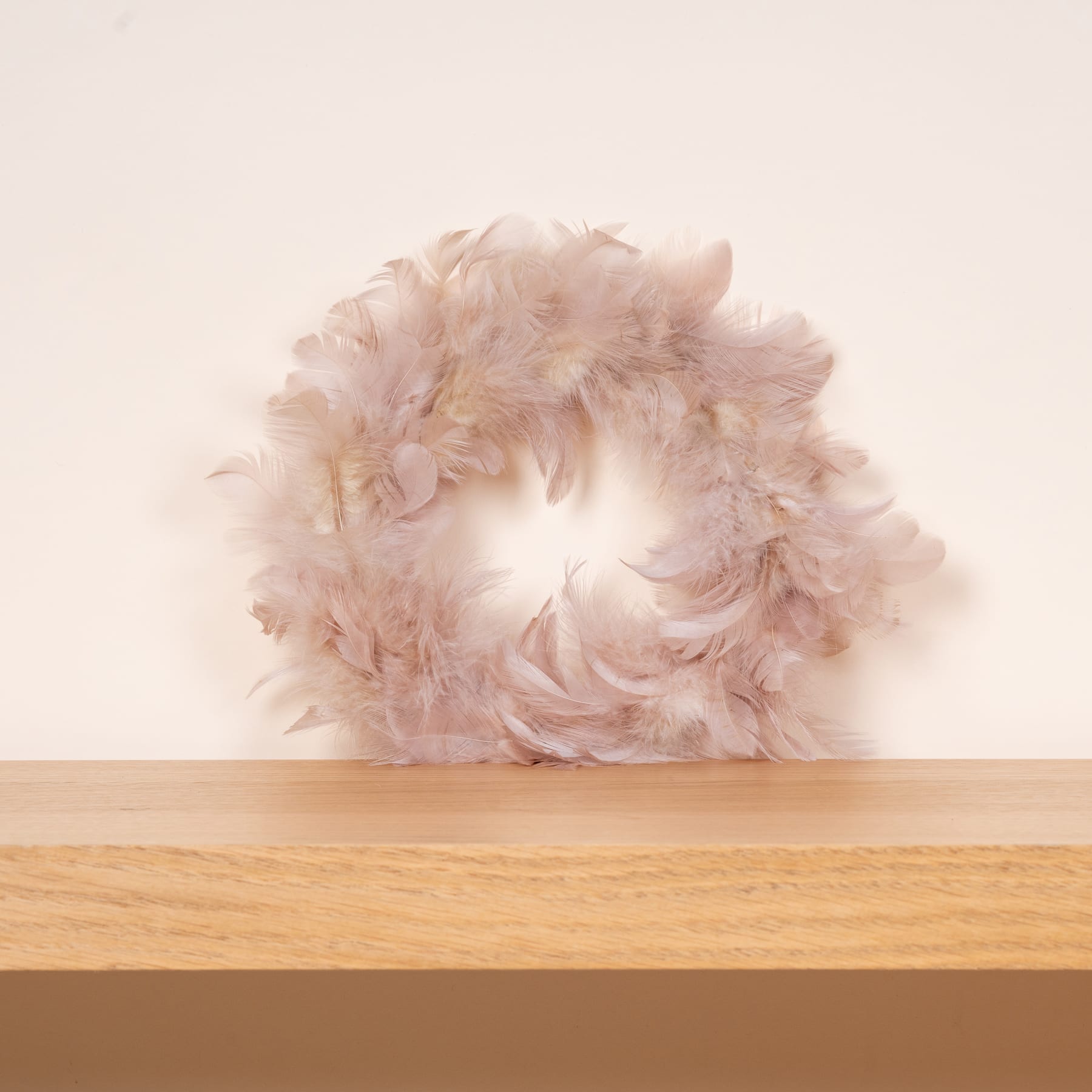 Ein flauschiger, blassrosa Federkranz sitzt auf einem Holzregal vor einem schlichten, hellen Hintergrund.