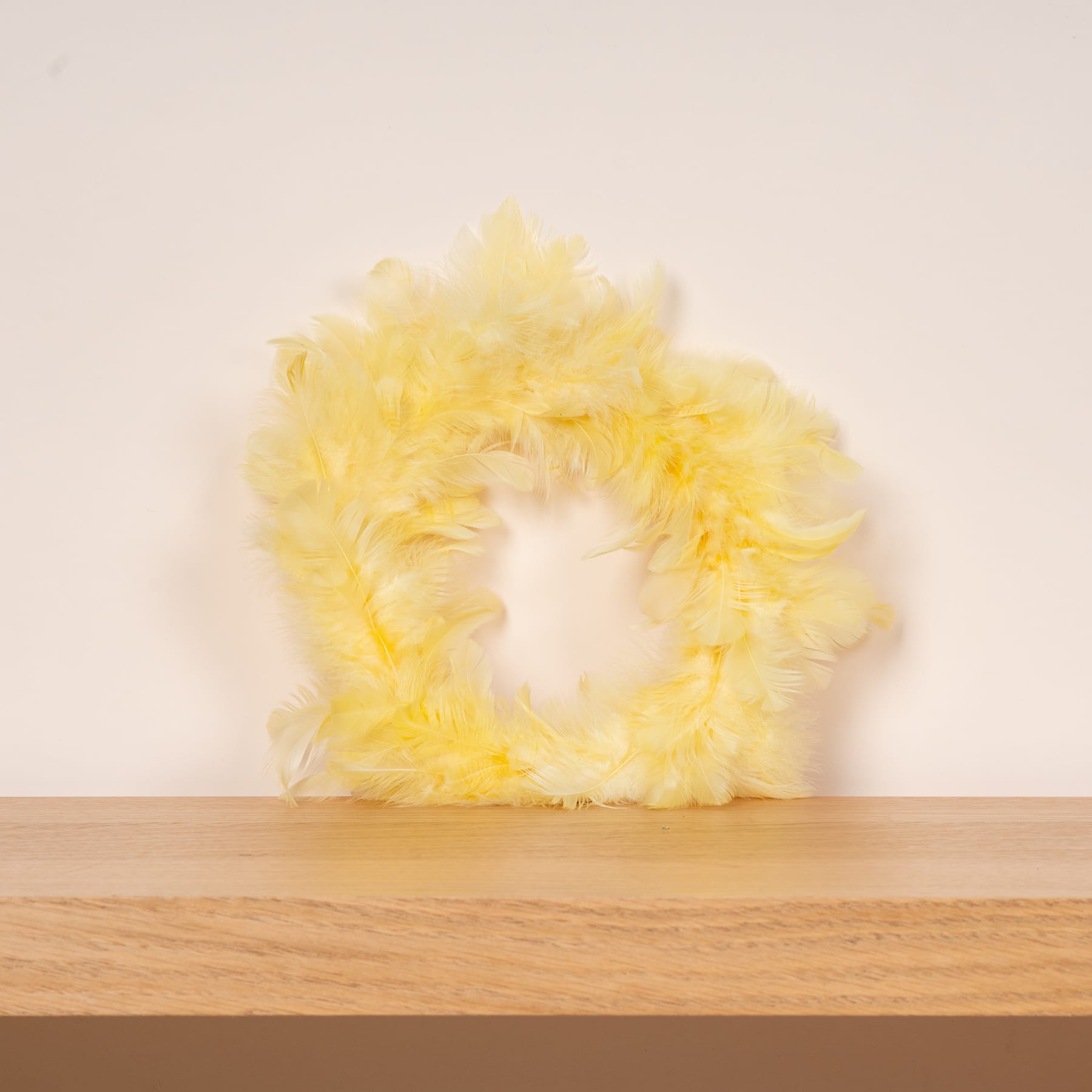 Ein flauschiger, gelber Federkranz liegt auf einer Holzoberfläche vor einem schlichten, hellen Hintergrund.