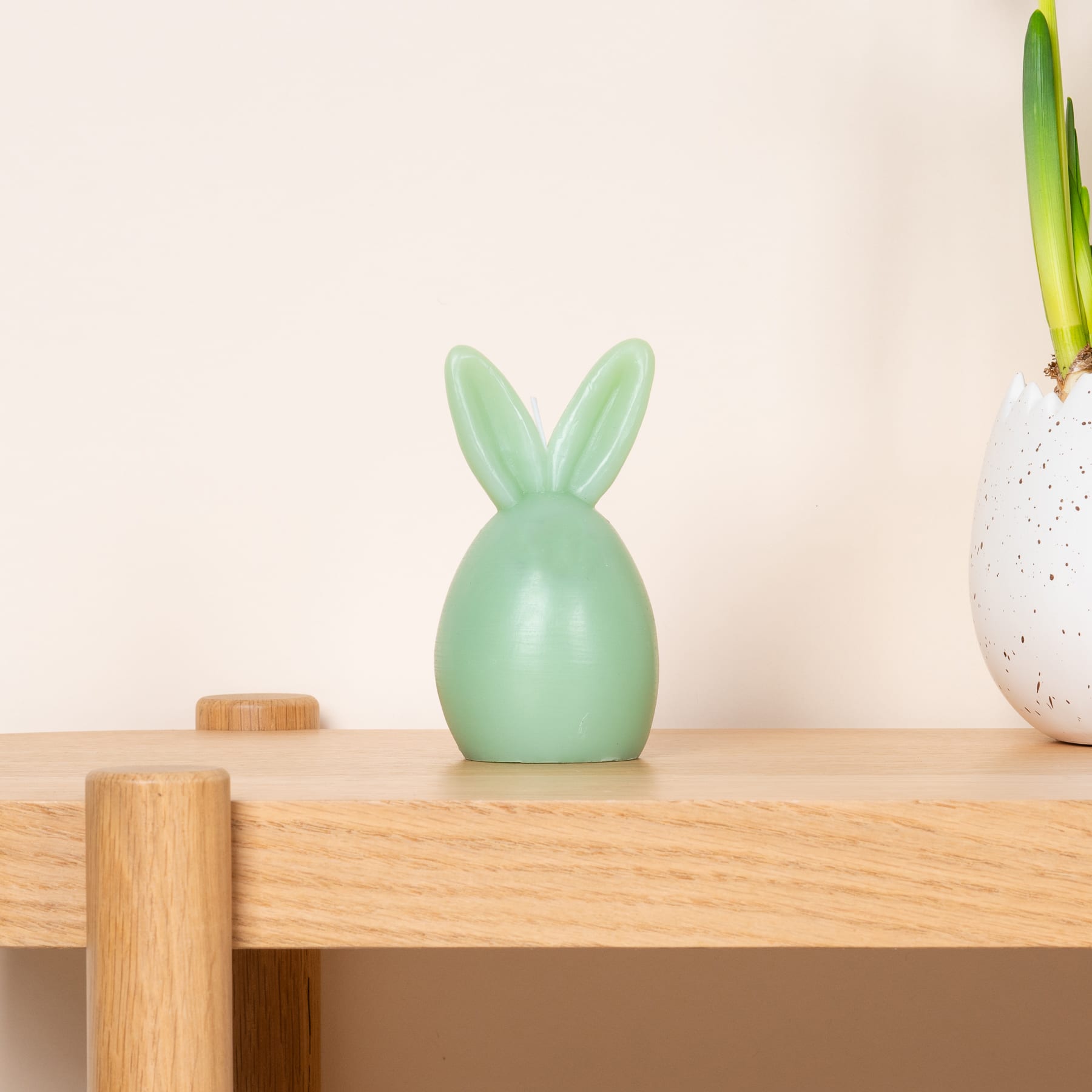 Auf einem Holztisch steht eine mintfarbene Kaninchenfigur aus Keramik, begleitet von einer Topfpflanze und einer gesprenkelten Keramikvase.